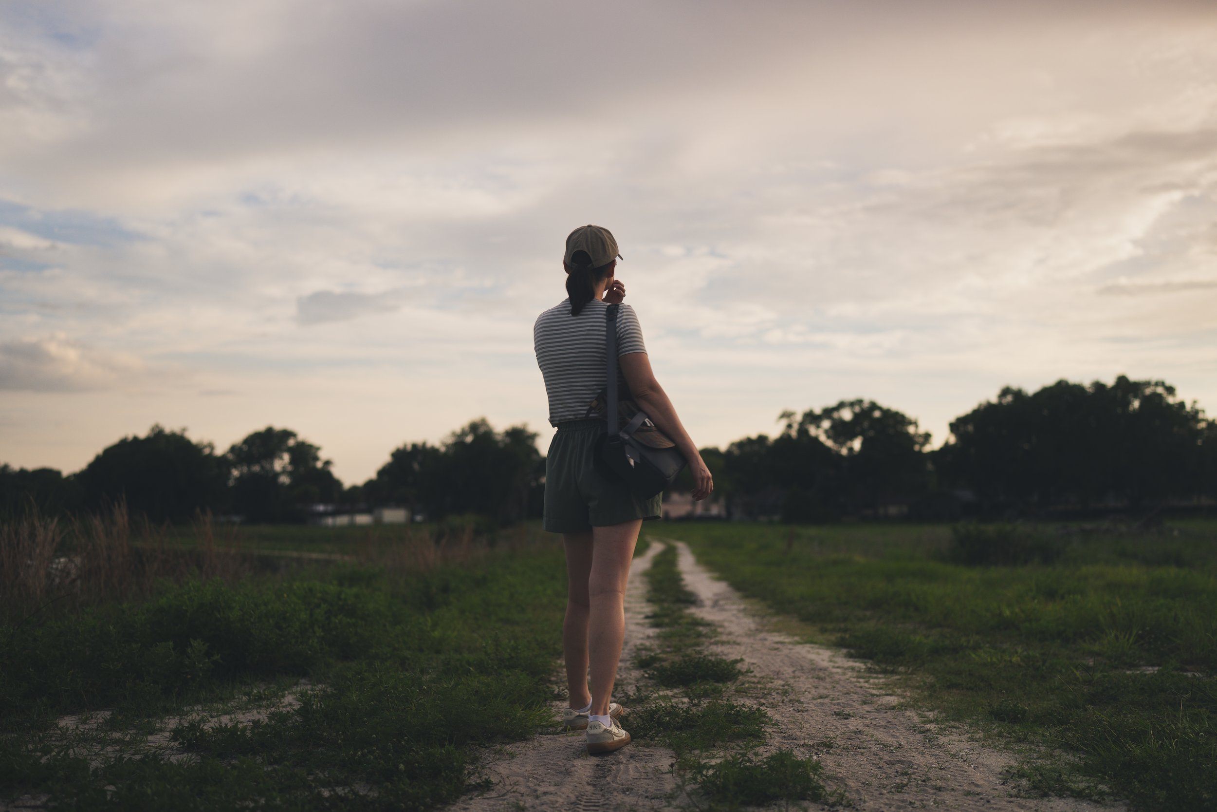 A woman in a striped t-shirt, wearing a cap, shorts, and sneakers, stands on a dirt path in a grassy area, looking into the distance during cloudy weather at dusk or dawn.
