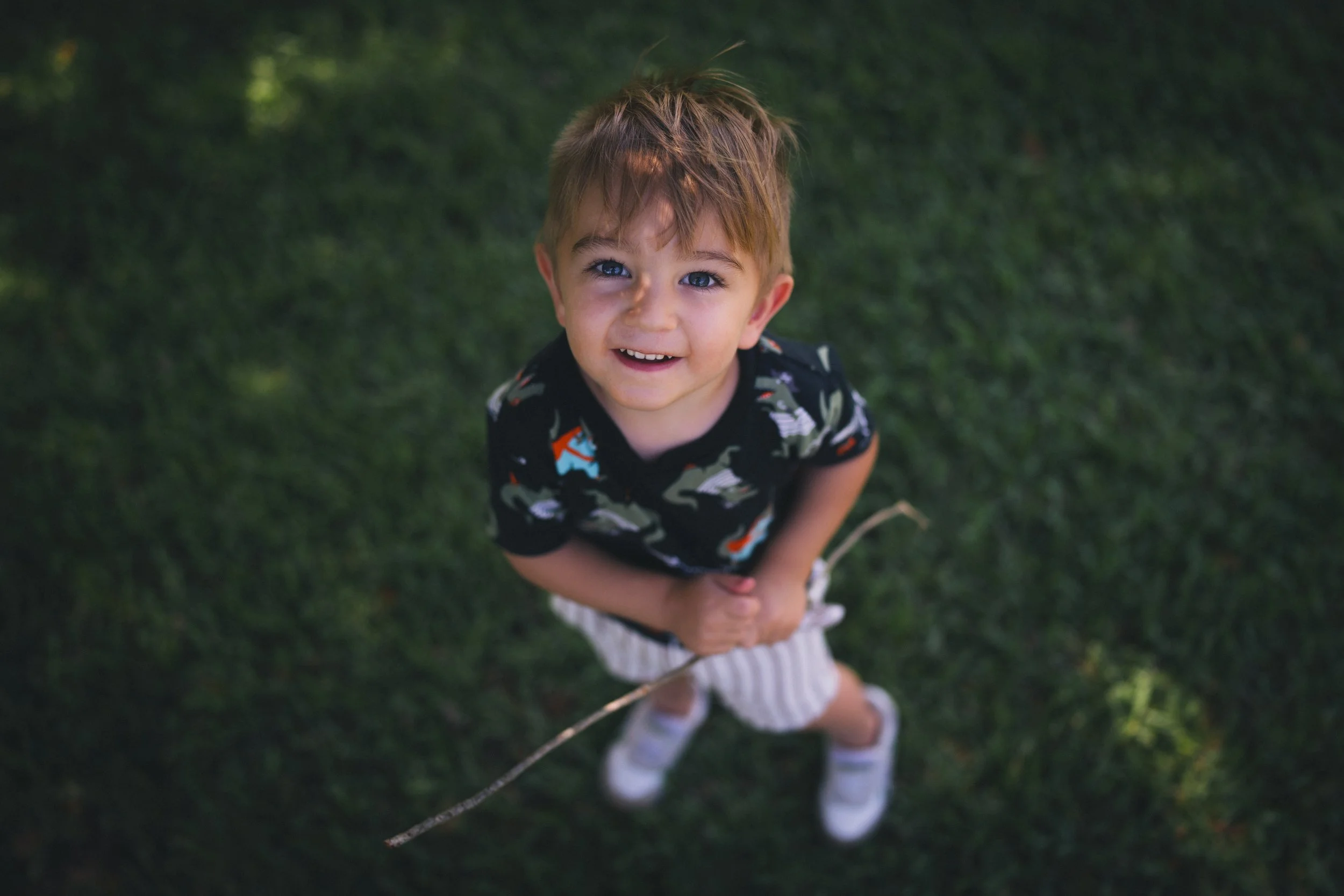 A young boy with light brown hair and blue eyes looking up at the camera, smiling, outdoors on grass.