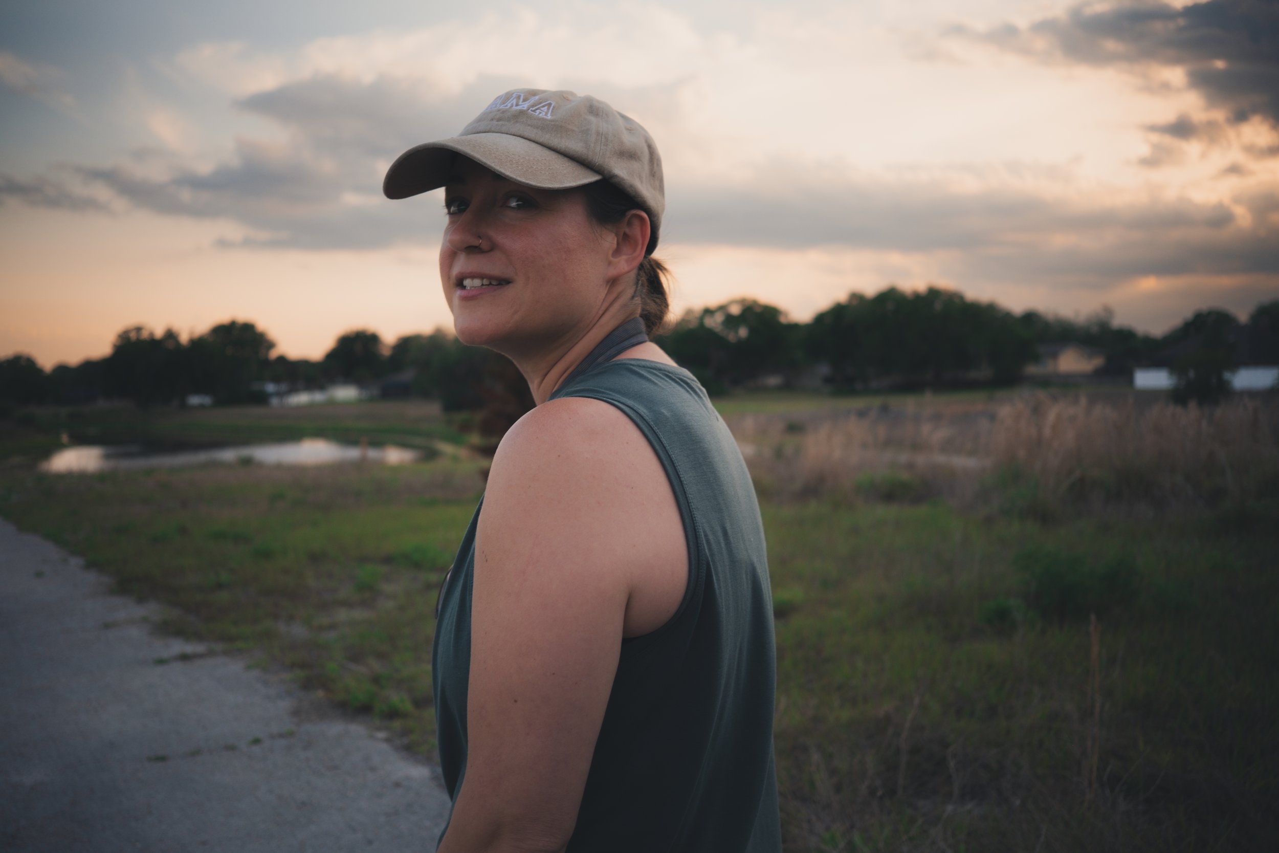 A woman with a beige cap and a nose ring looking over her shoulder at sunset outdoors, with trees and a cloudy sky in the background.