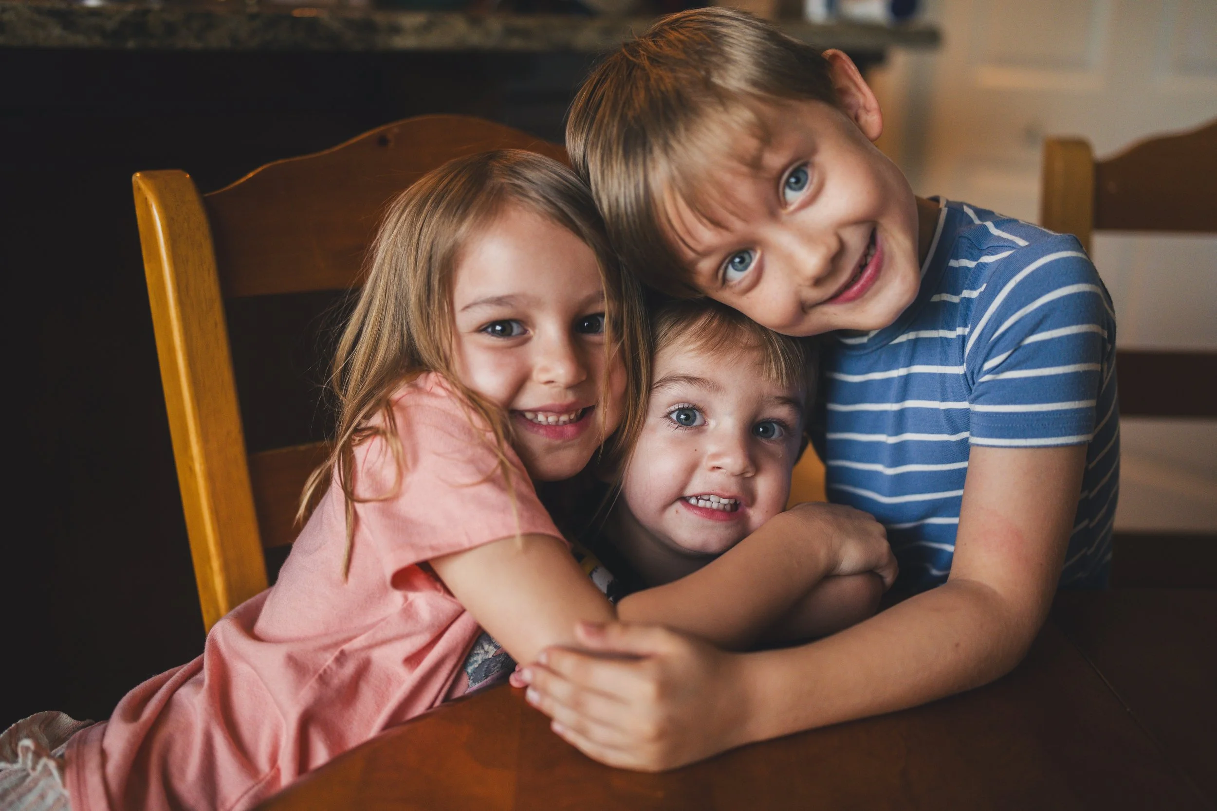 Three children, two boys and a girl, smiling and hugging each other at a wooden dining table in a warmly lit room.