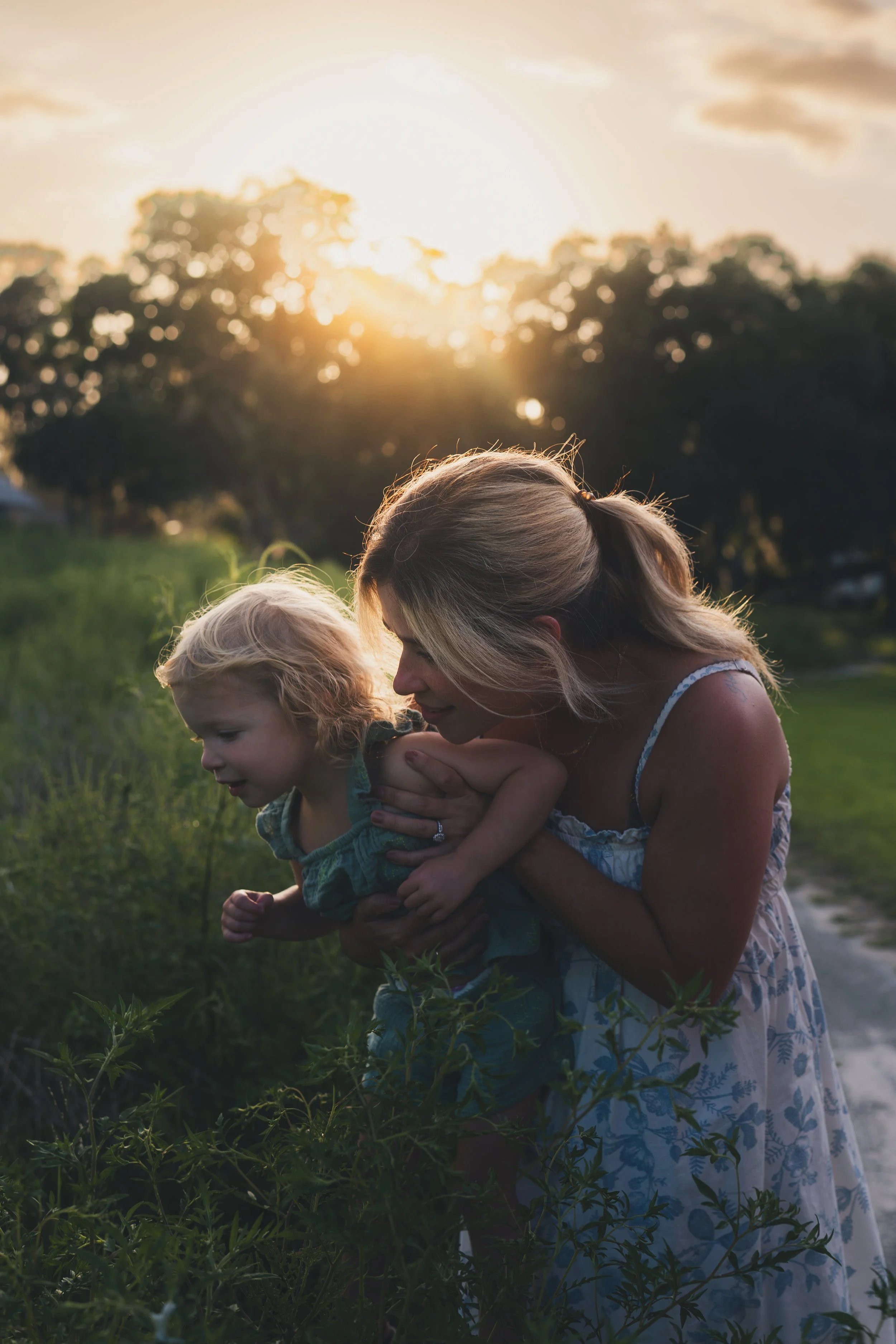 A woman and a young child are outdoors during sunset, surrounded by green plants, with the woman holding the child close and they are smiling at each other.
