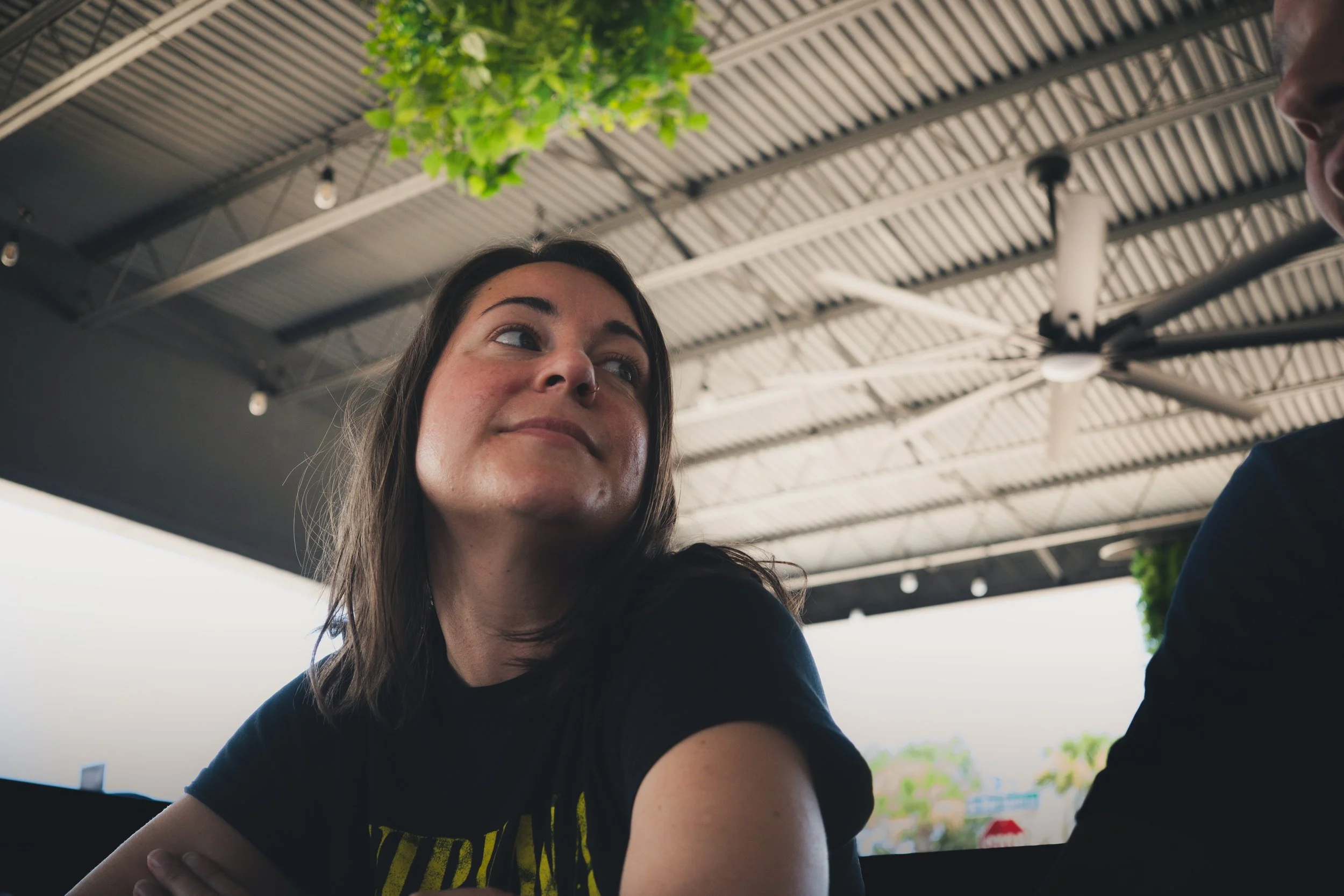 A woman with brown hair and a black t-shirt with yellow text, sitting under a metal roof with hanging lights and green plants, looking to her right.