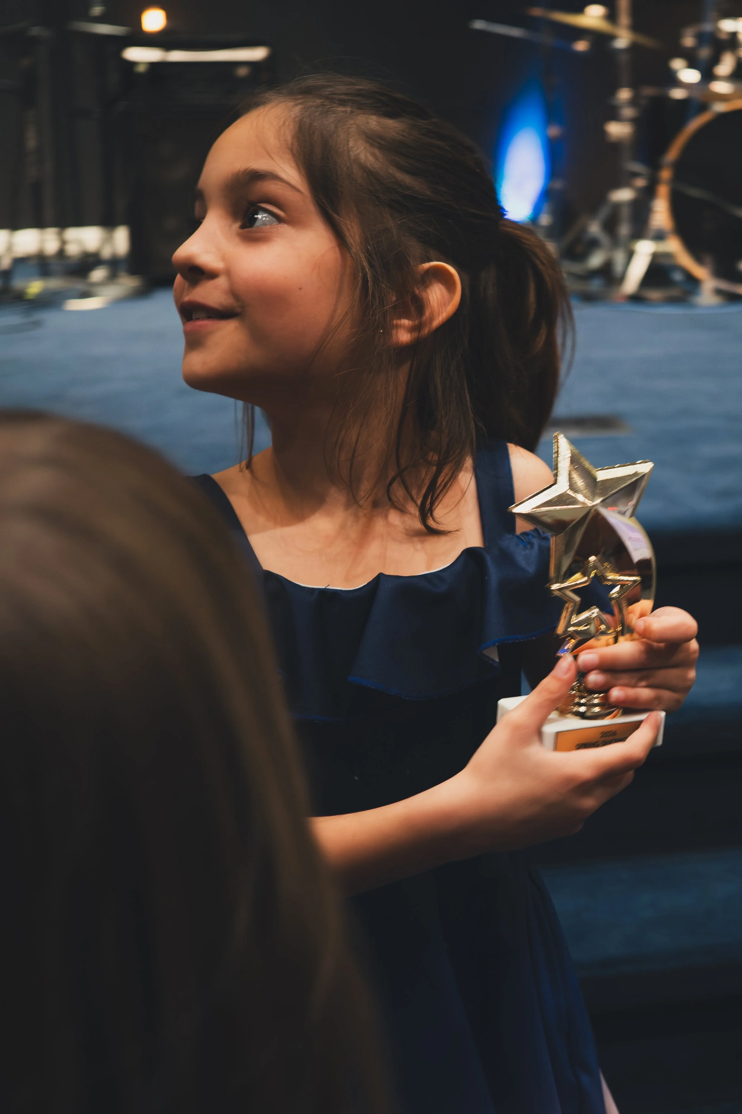 A young girl receiving an award or trophy at an event, holding a star-shaped trophy made of gold, with people and event equipment in the background.