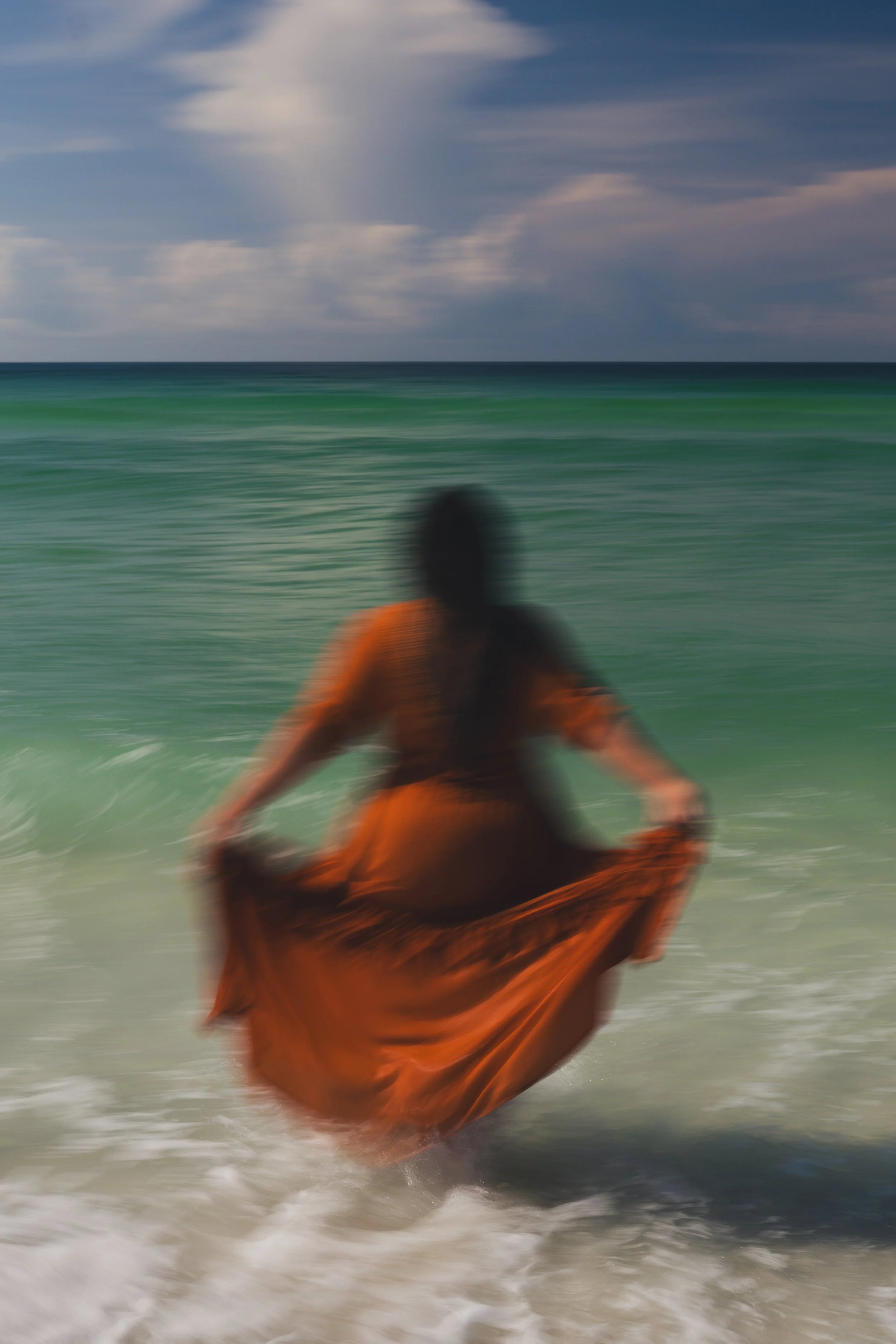 Blurred image of a person wearing an orange garment sitting on the beach with ocean waves in the background and partly cloudy sky.
