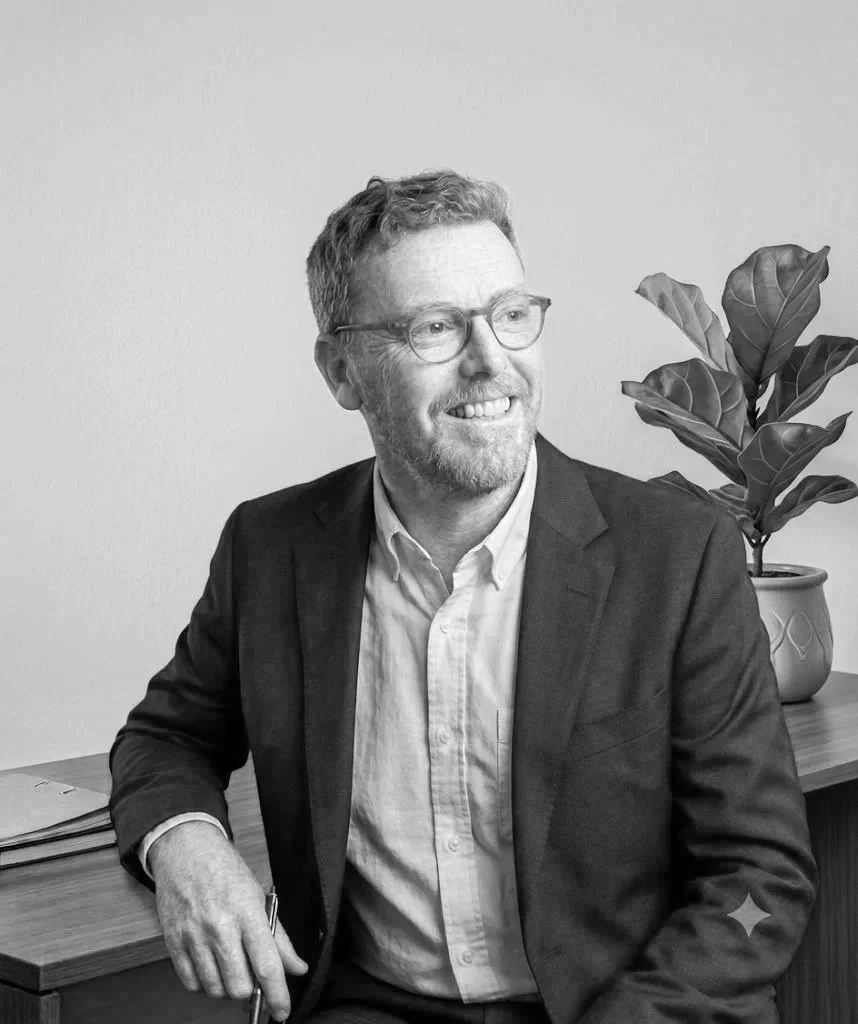 A black-and-white photo of a man with glasses, facial hair, and a friendly smile, sitting at a wooden desk with a potted plant behind him.