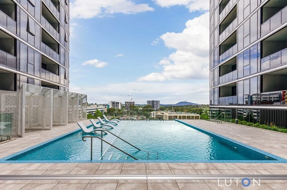 Empty rooftop swimming pool between two modern high-rise buildings with lounge chairs and city skyline in the background.