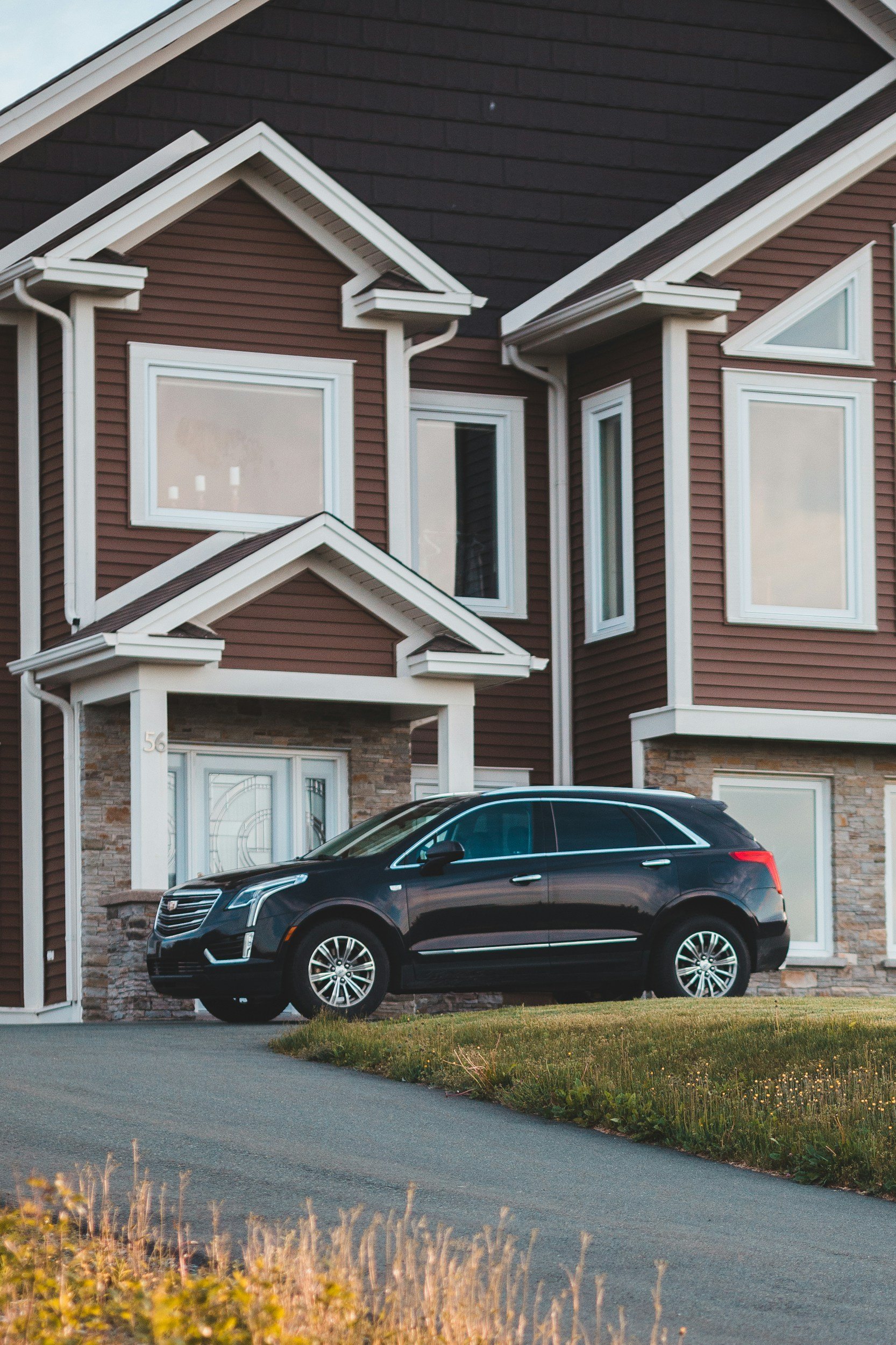 Front view of a large, modern house with dark and red siding, multiple gable roofs, and white trim. A black luxury SUV is parked in the driveway in front of the house.