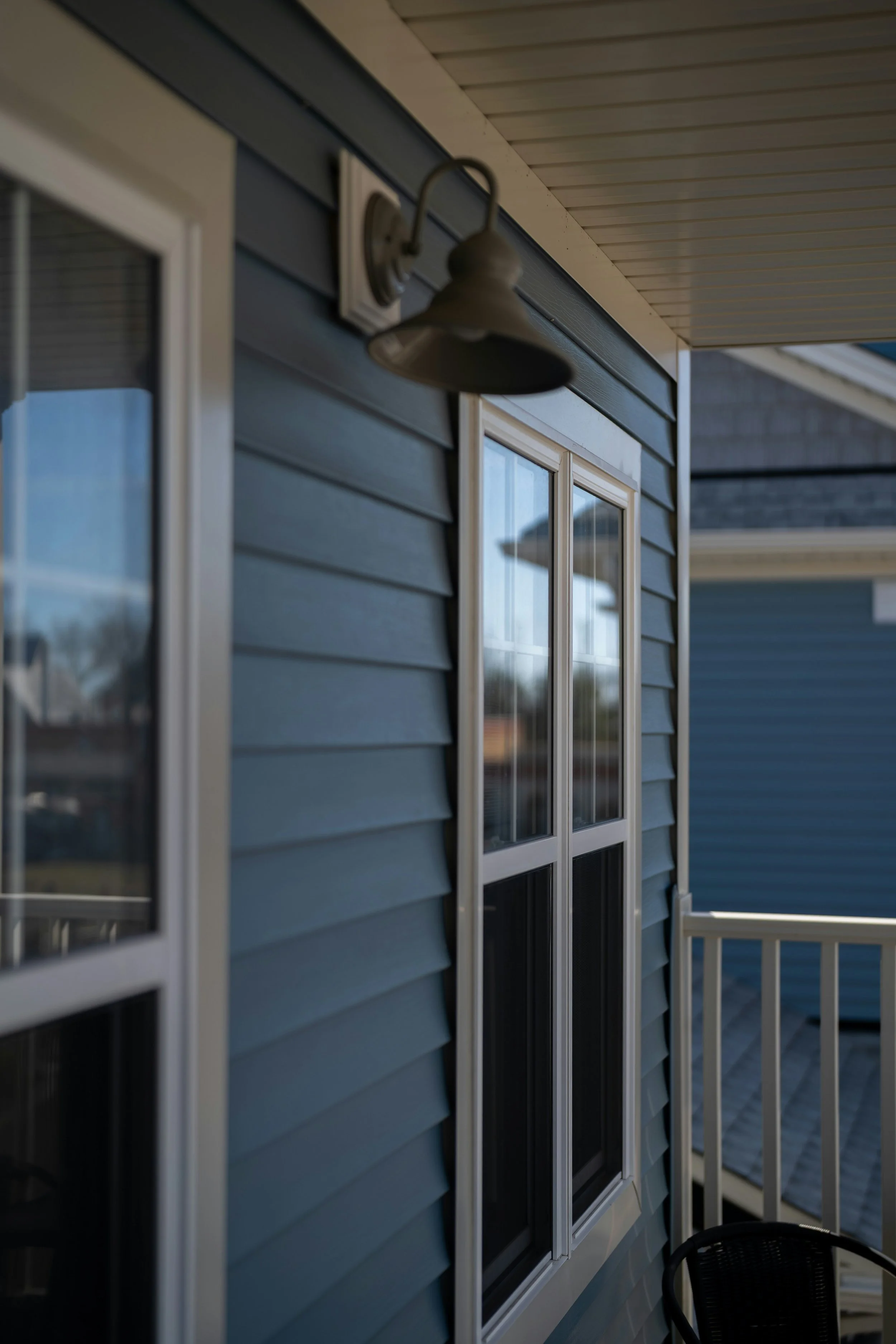 A shot of a house exterior showing two windows, a light fixture, and part of a porch railing. The house has blue siding and a white-trimmed window.