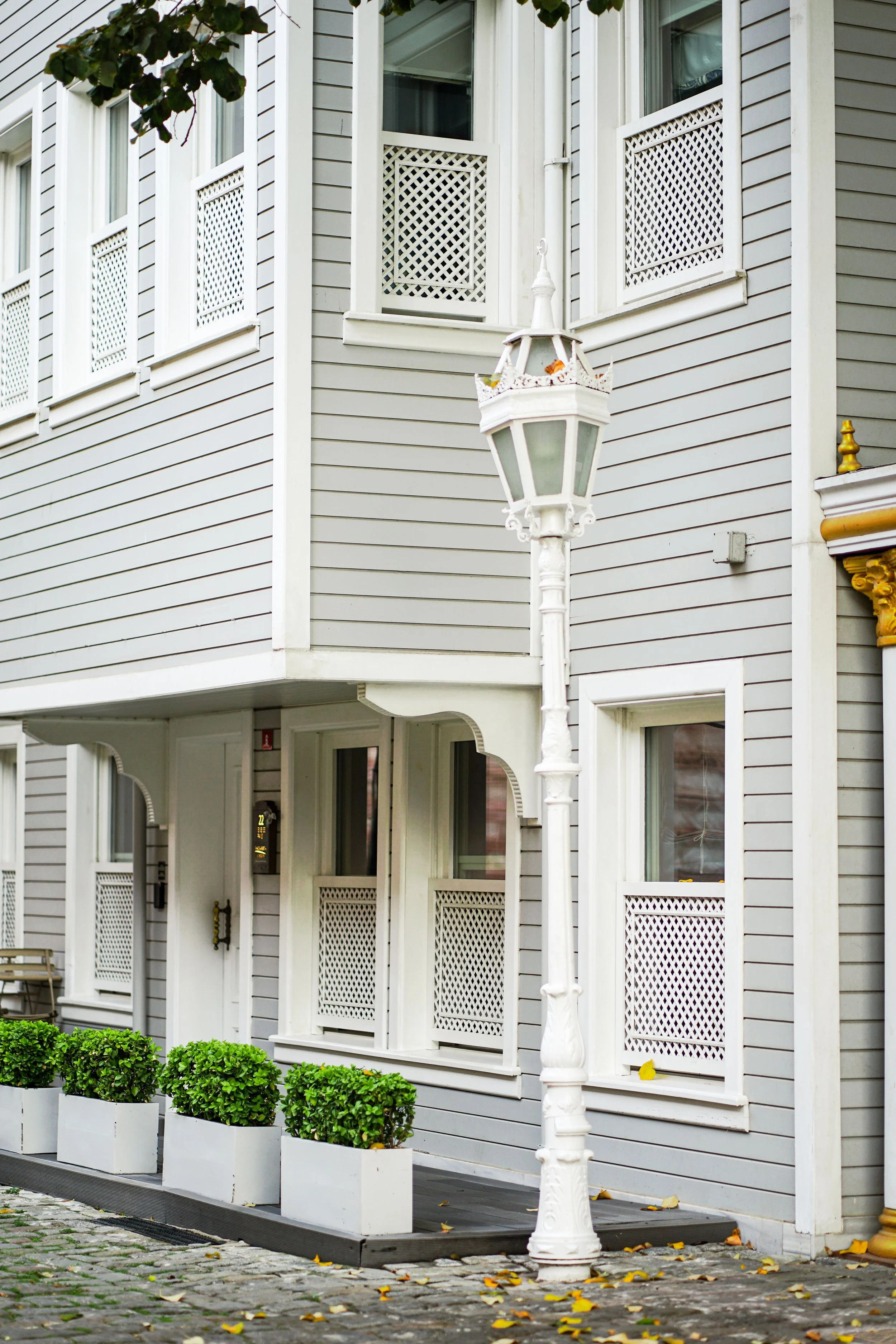 Exterior view of a multi-story residential building with gray siding, white window frames, decorative lattice panels, and a vintage white street lamp. Potted bushes line the sidewalk, with fallen leaves scattered on the cobblestone pavement.