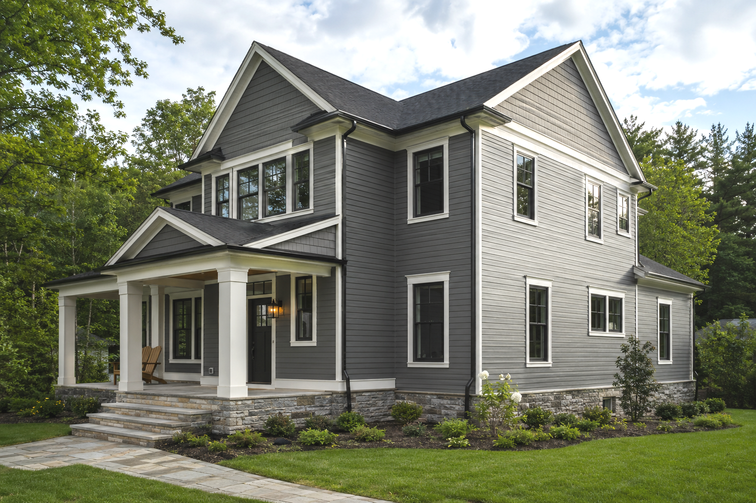 A gray, two-story house with white trim and a stone foundation, surrounded by green grass and trees, under a partly cloudy sky.