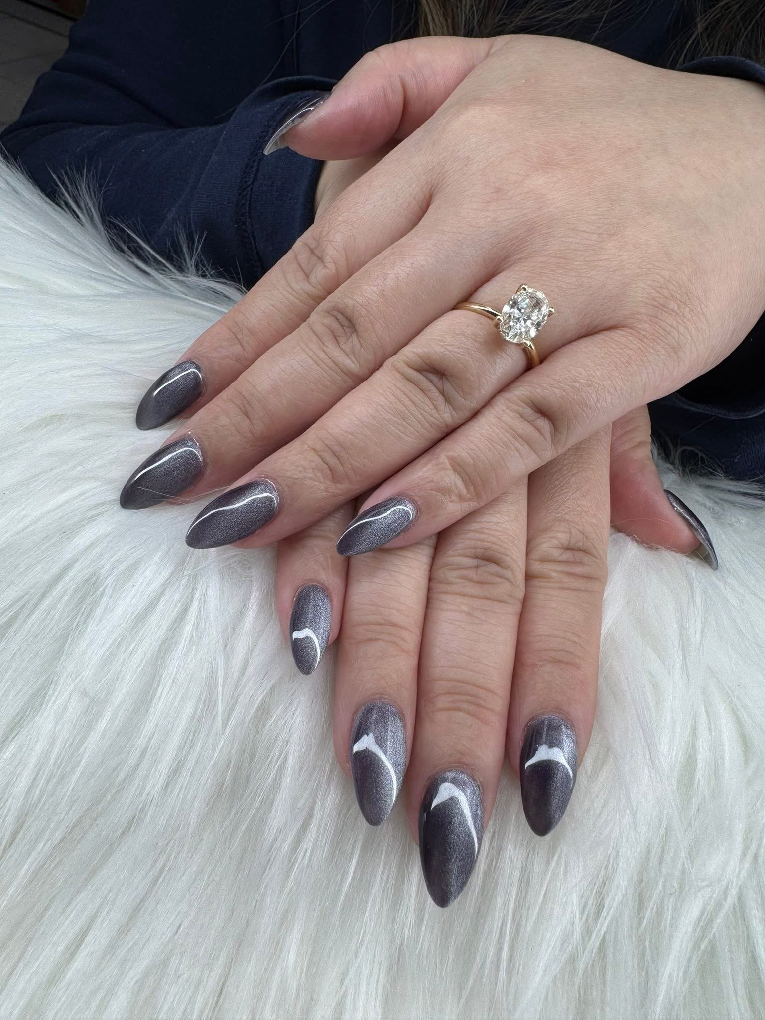 Close-up of hands with manicured nails and an engagement ring with a large diamond on the ring finger, resting on a white furry surface.