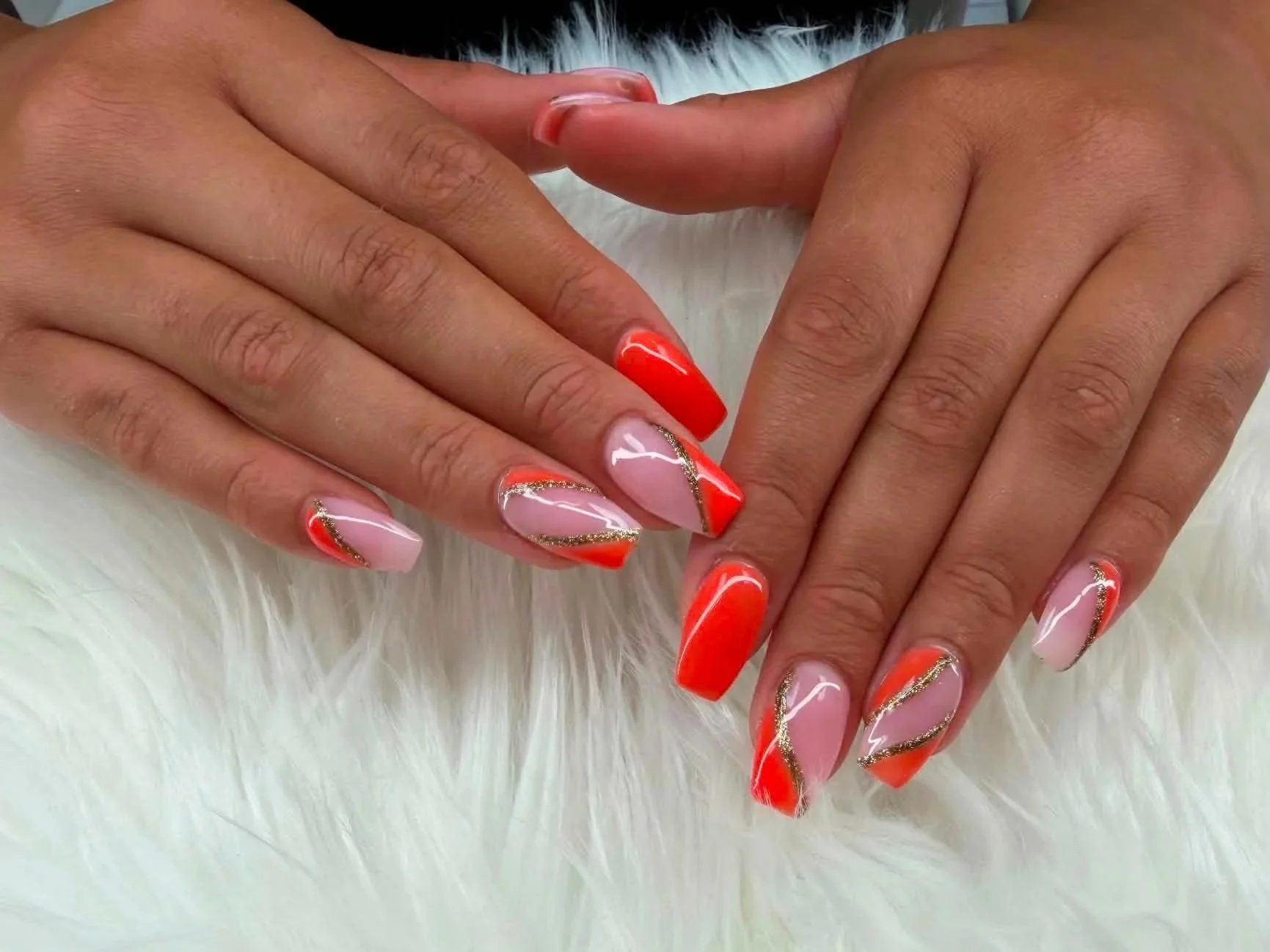Manicured nails with orange, pink, and gold glitter design, resting on fluffy white surface.