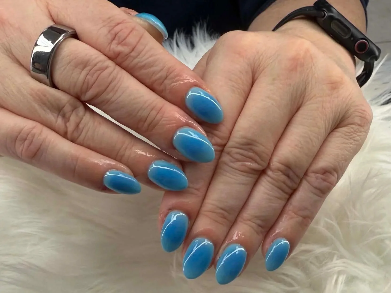 Close-up of a person's hands with blue painted nails, multiple rings, and a black wristband, resting on a white furry surface.