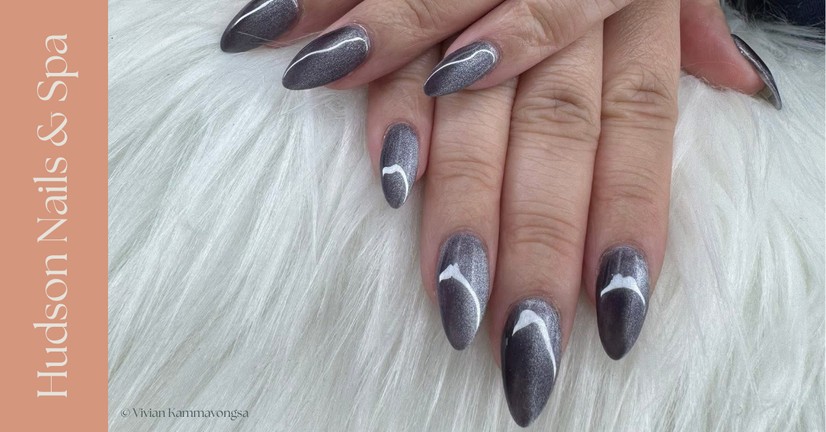 Close-up of a hand with long oval-shaped nails painted in metallic dark gray with white curved stripe accents, resting on white fur background. The side of the image features a pale pink vertical banner with the text 'Hudson Nails & Spa' in white.