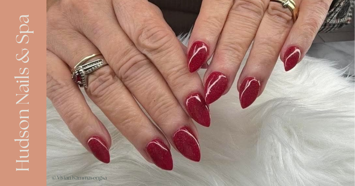 Close-up of hands with red, almond-shaped manicured nails resting on a white furry surface, wearing rings.