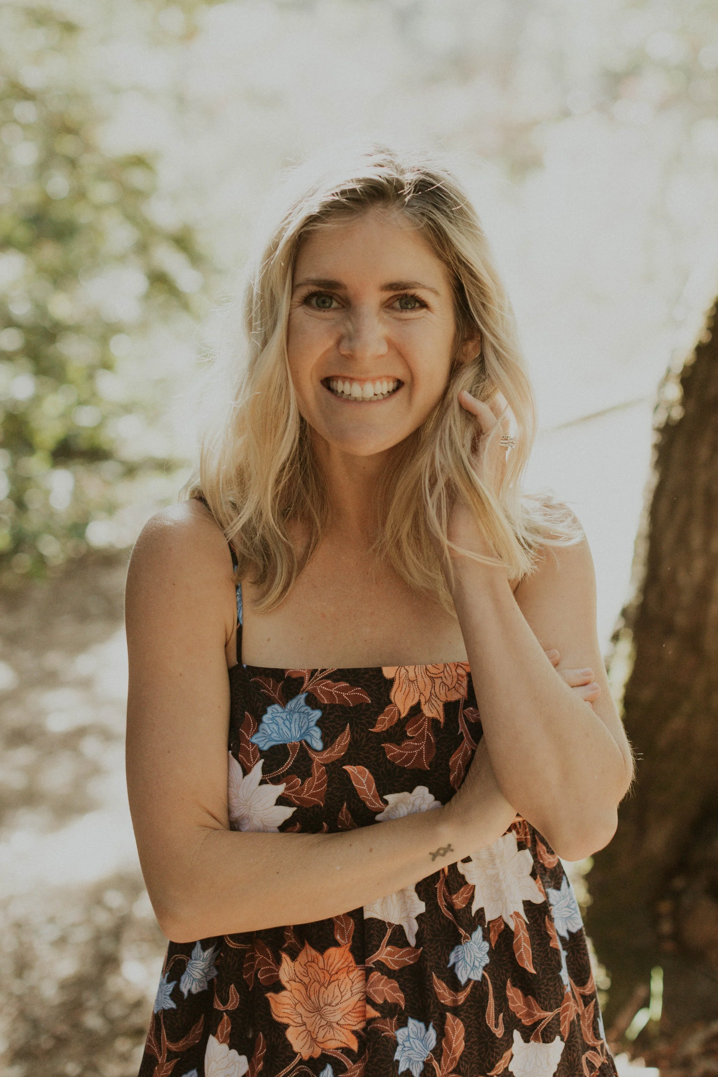 A woman with blonde hair smiling outdoors, wearing a sleeveless dress with a floral pattern, standing with one hand touching her face.