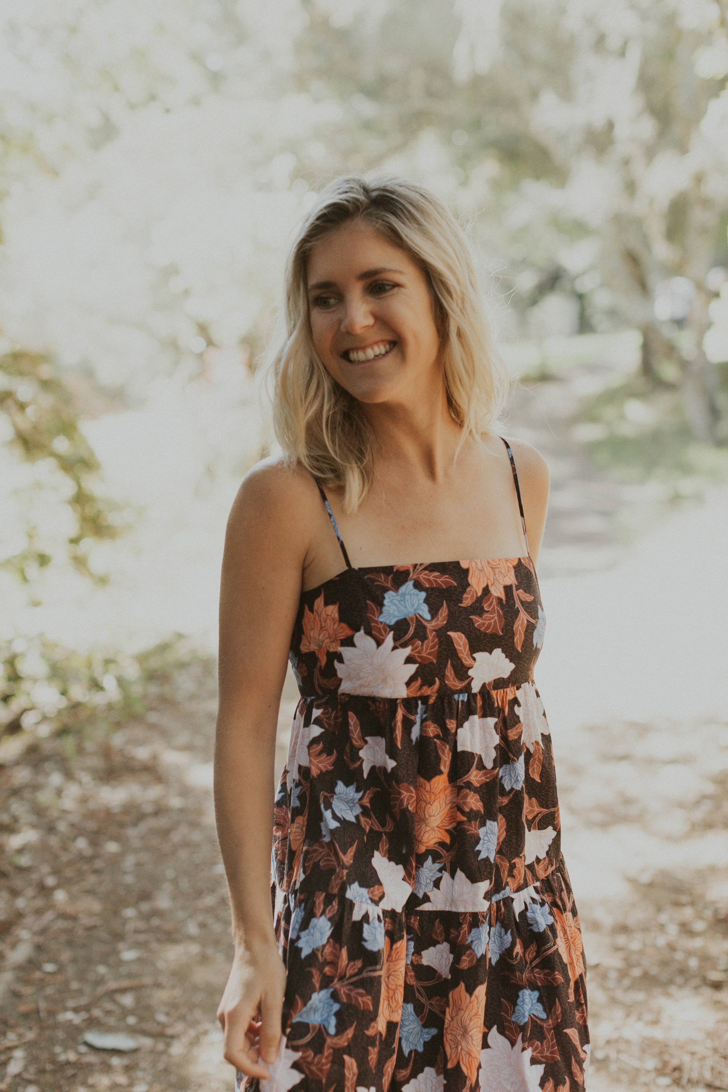 A woman with blonde hair in a floral dress standing outdoors on a dirt path surrounded by trees.
