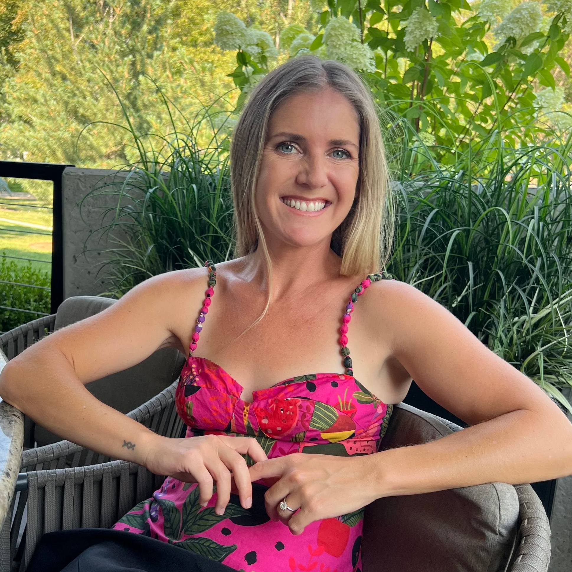 A woman with blonde hair and blue eyes smiling while sitting outdoors on a patio, wearing a colorful floral dress and beaded necklace, surrounded by greenery and plants.