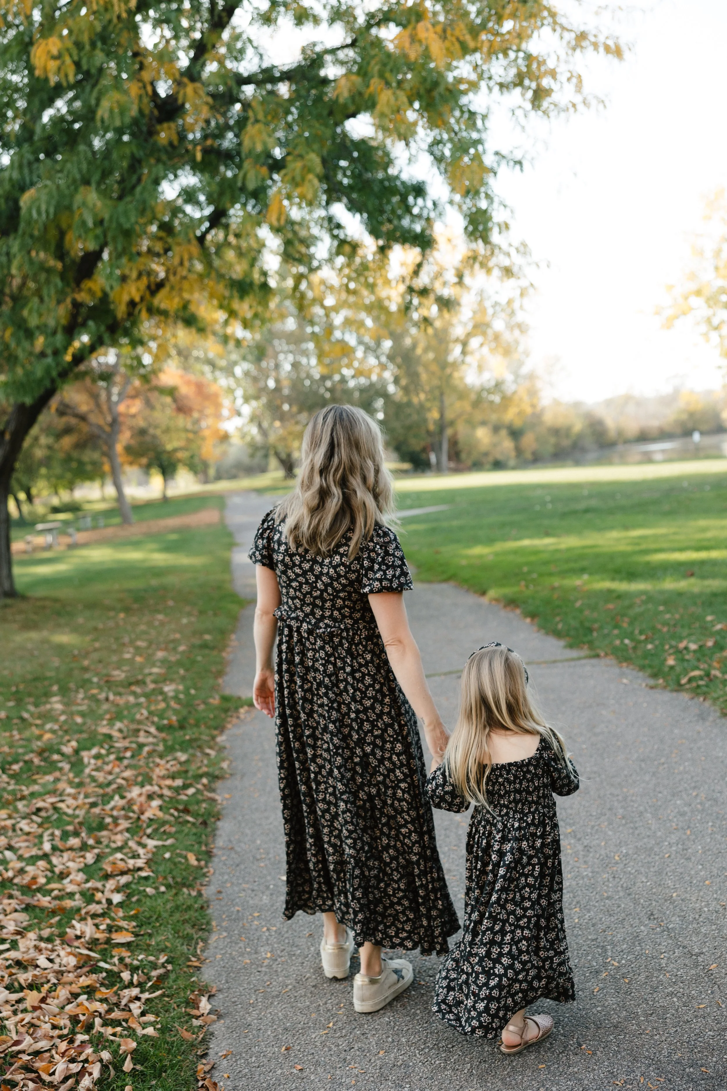 A woman and a young girl walking hand-in-hand on a paved pathway in a park during autumn. They are wearing matching black dresses with white floral patterns. The park has green grass, falling leaves, and trees with green and yellow foliage. The woman is wearing white sneakers, and the girl is wearing pink sandals.