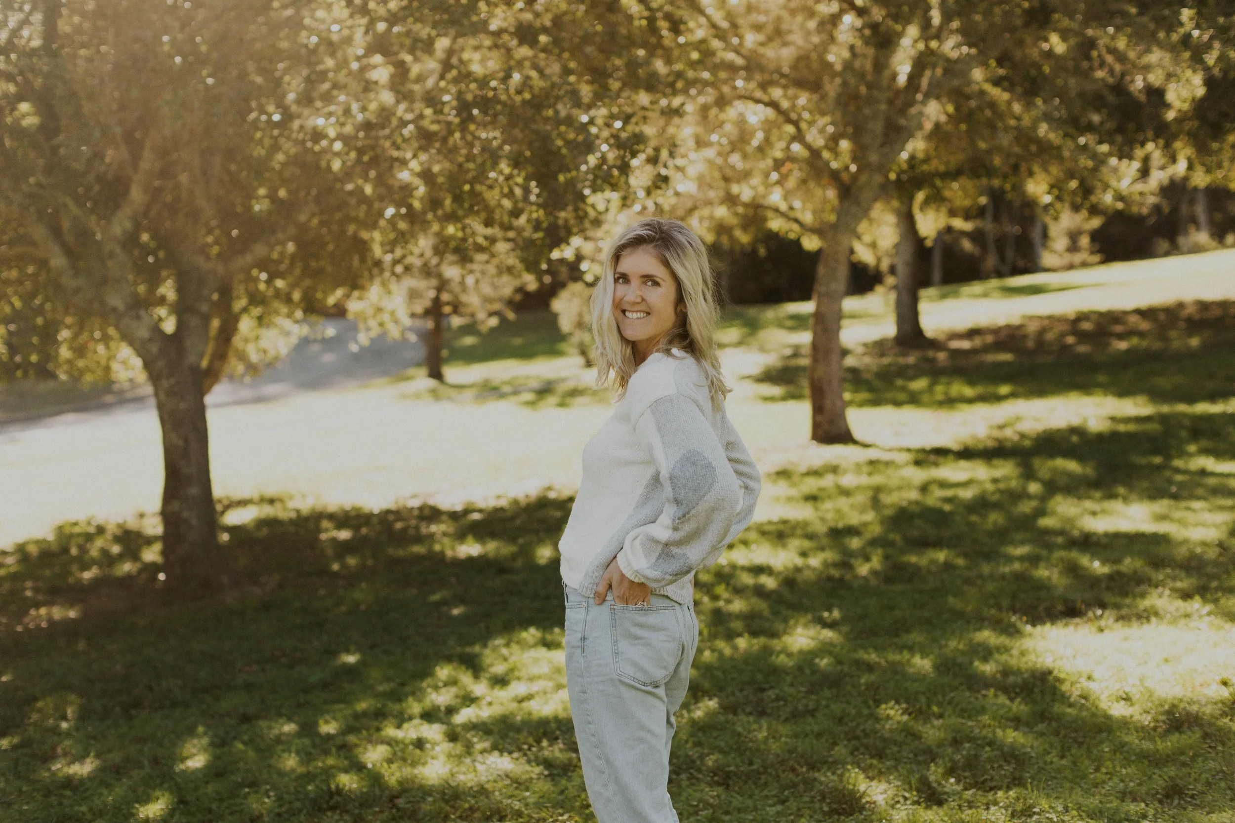 A young woman with blonde hair smiling and looking over her shoulder in a park with trees and sunlight filtering through the leaves.