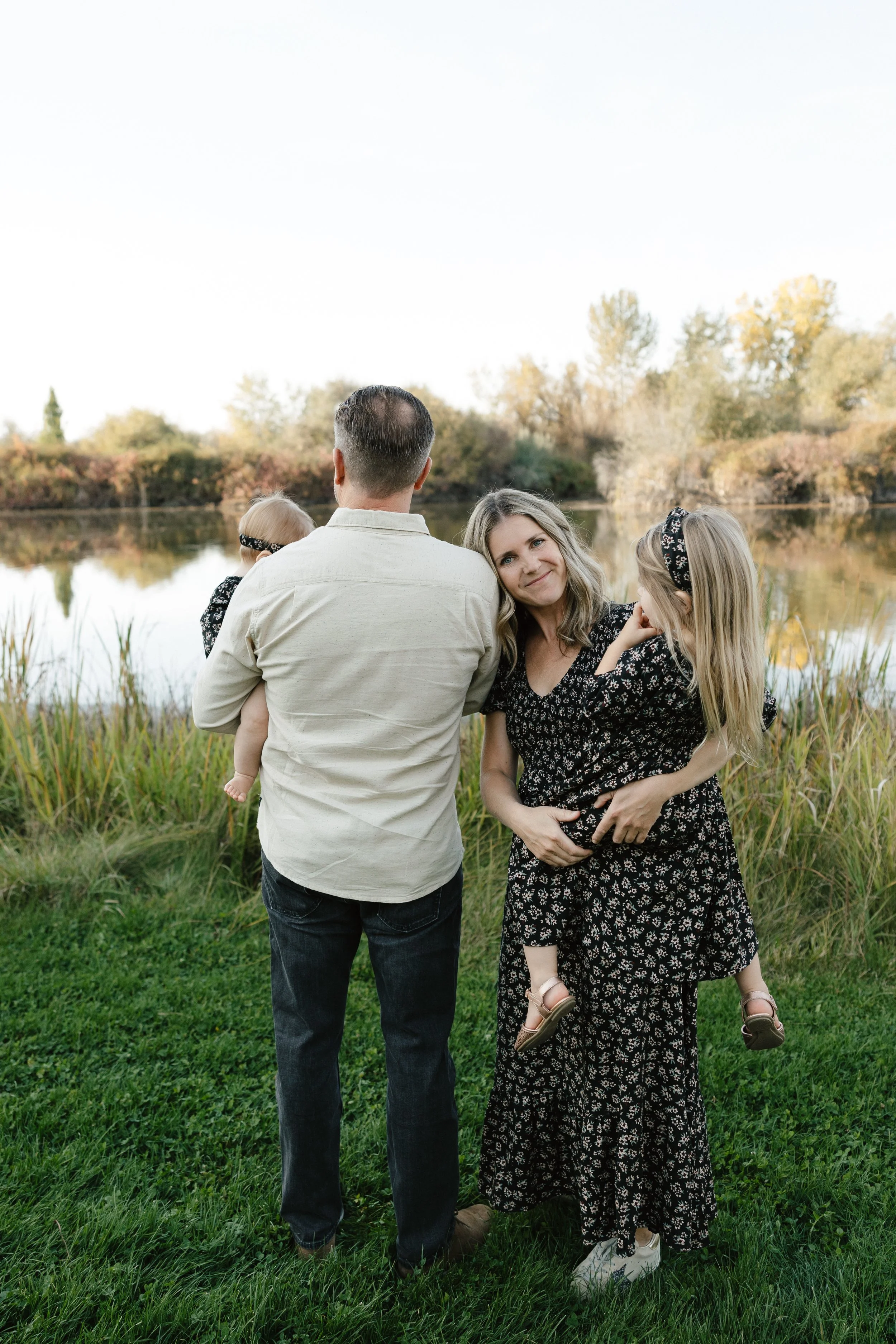 A family of four standing by a river during autumn, with trees in fall colors in the background. The man is holding a young girl, and the woman is holding a young girl in her arms, all facing away from the camera and the woman smiling at the camera.