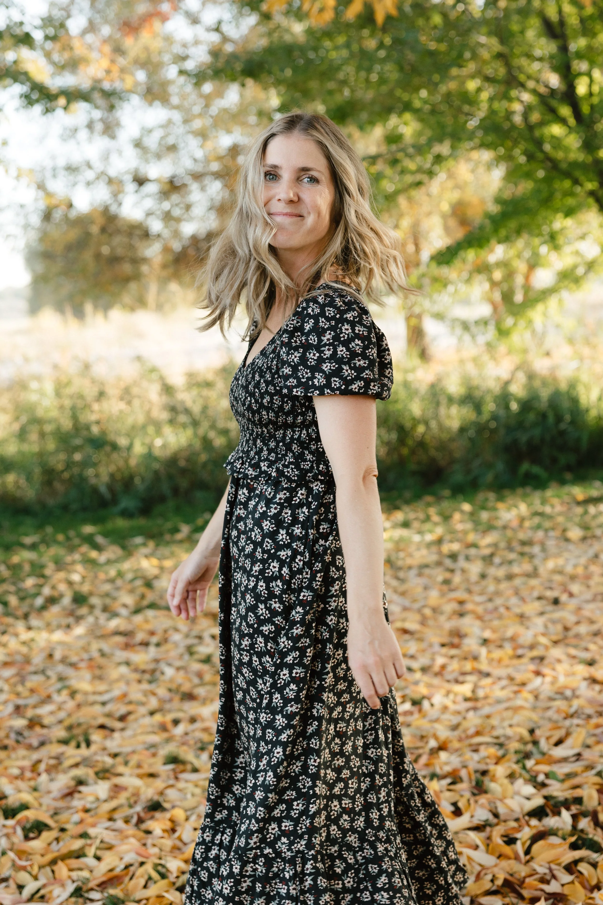 A woman with wavy blonde hair wearing a black dress with small white floral patterns, standing outdoors among yellow and green autumn leaves.