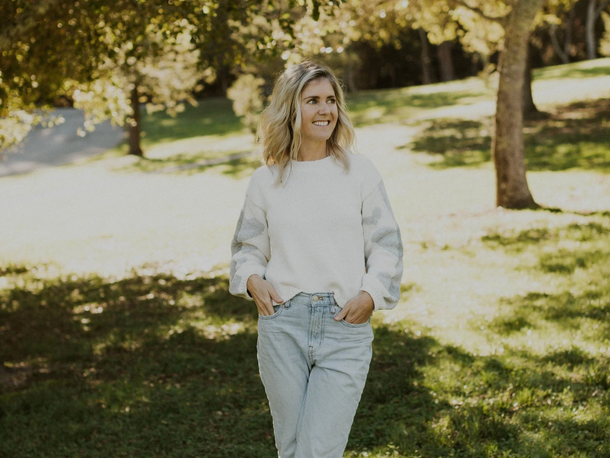 A woman with blonde, wavy hair wearing a white sweater and light blue jeans standing outdoors in a park with trees and sunlight.