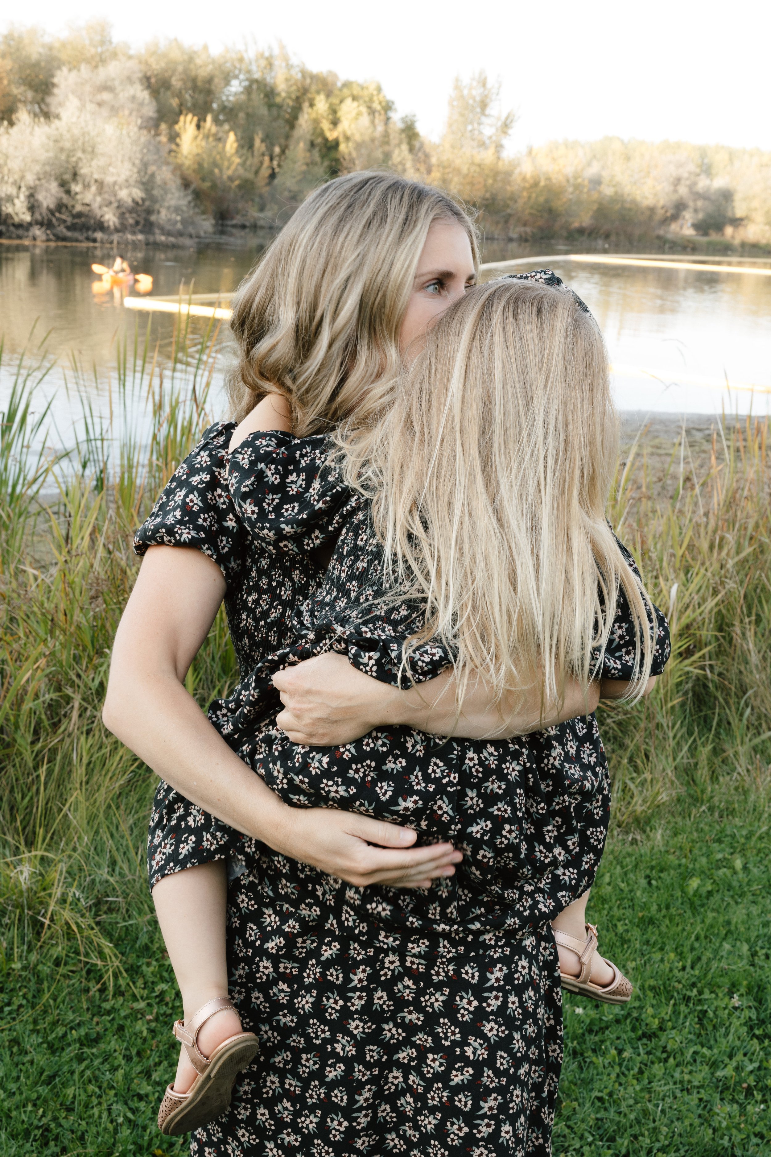 A woman hugging a young girl outdoors near a lake with ducks swimming and trees in the background.