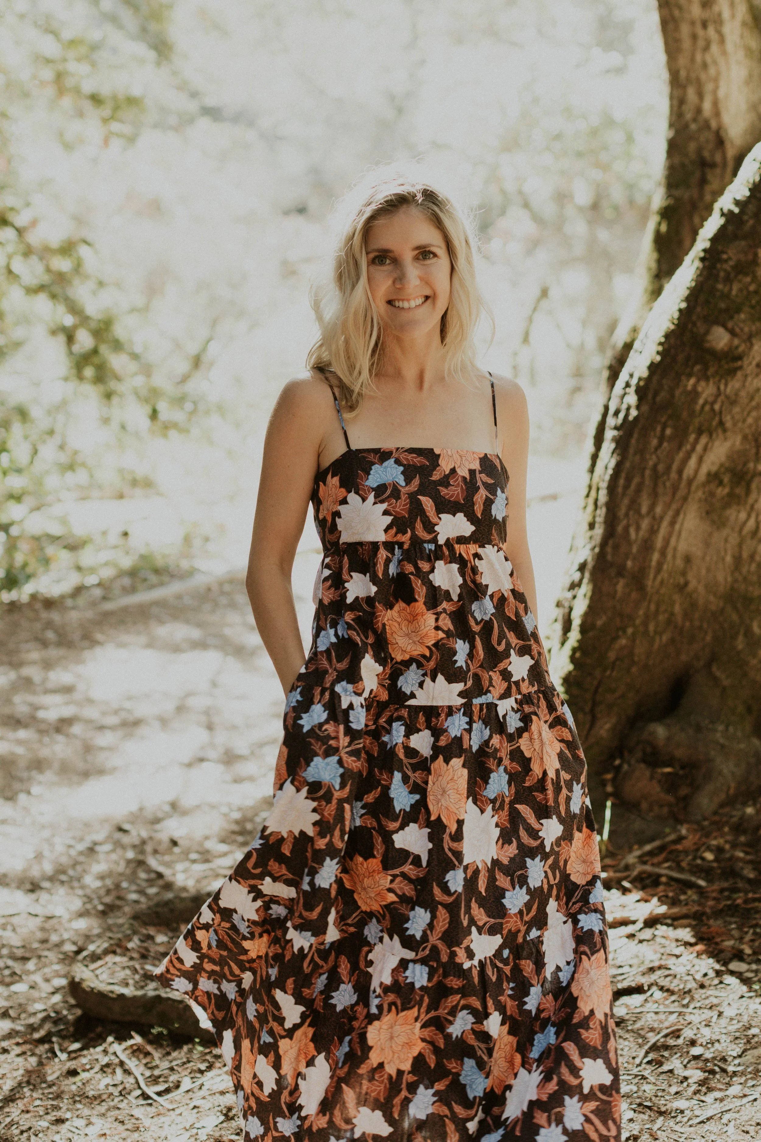 A woman smiling outdoors near a tree, wearing a floral dress with black, white, blue, and orange flowers.