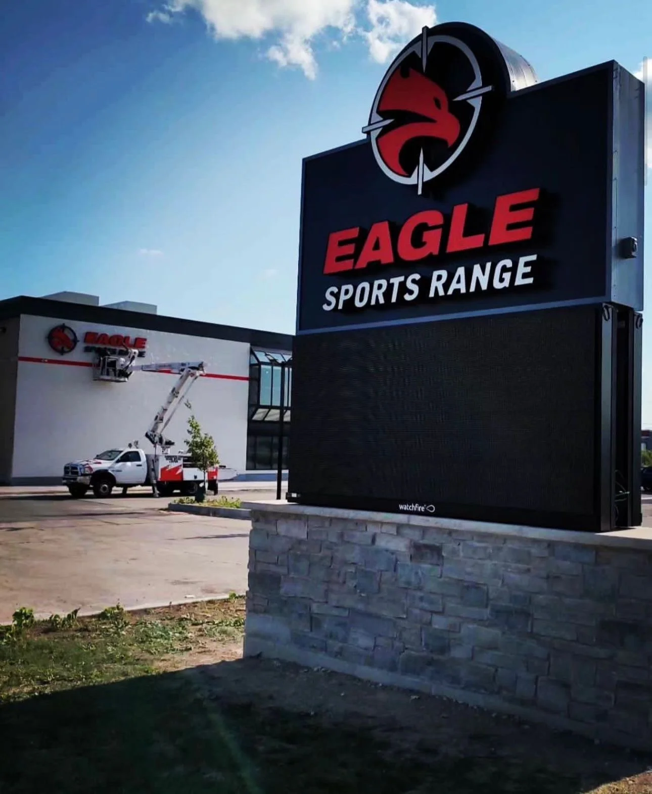 Eagle Sports Range sign with a logo of a red eagle head in a target circle, and a building in the background with a cherry picker and vehicle.
