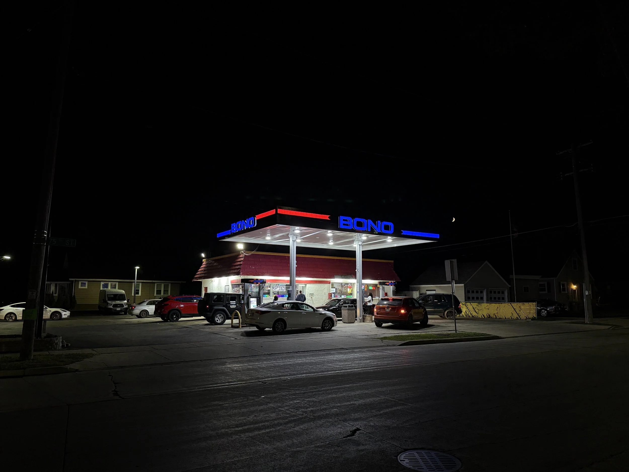 A nighttime view of a gas station with a red and white canopy, illuminated with bright lights, and the name 'BONO' displayed in blue and red on the signage. Several cars are parked nearby, and the surrounding area includes small buildings and streetlights.