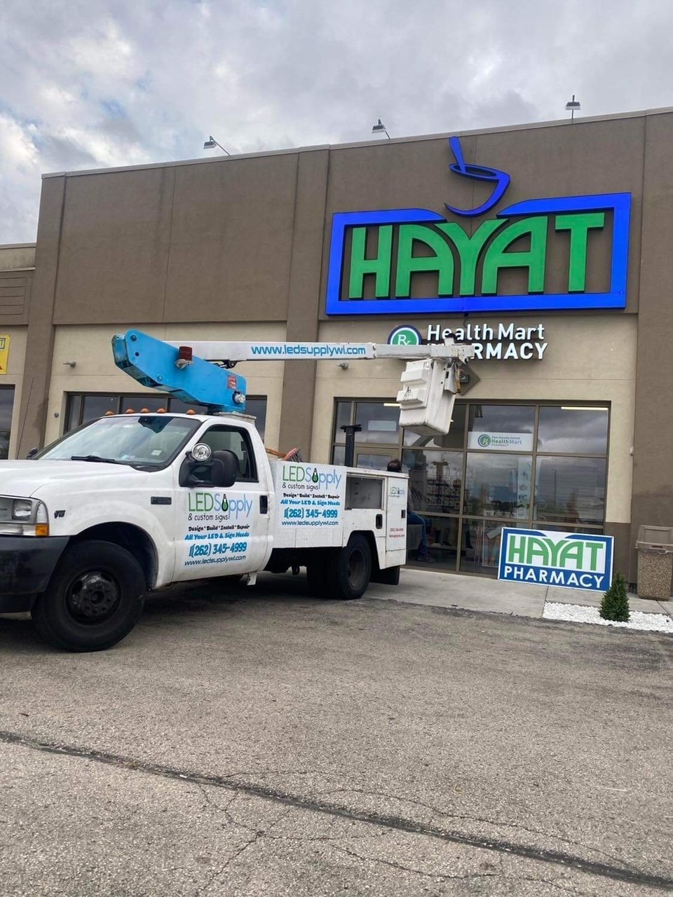 A truck with a bucket lift parked in front of a pharmacy named Hayat. The pharmacy has a sign with its name in green and blue, and there is an additional sign on the ground near the entrance. The truck is branded with LED Supply advertisements.