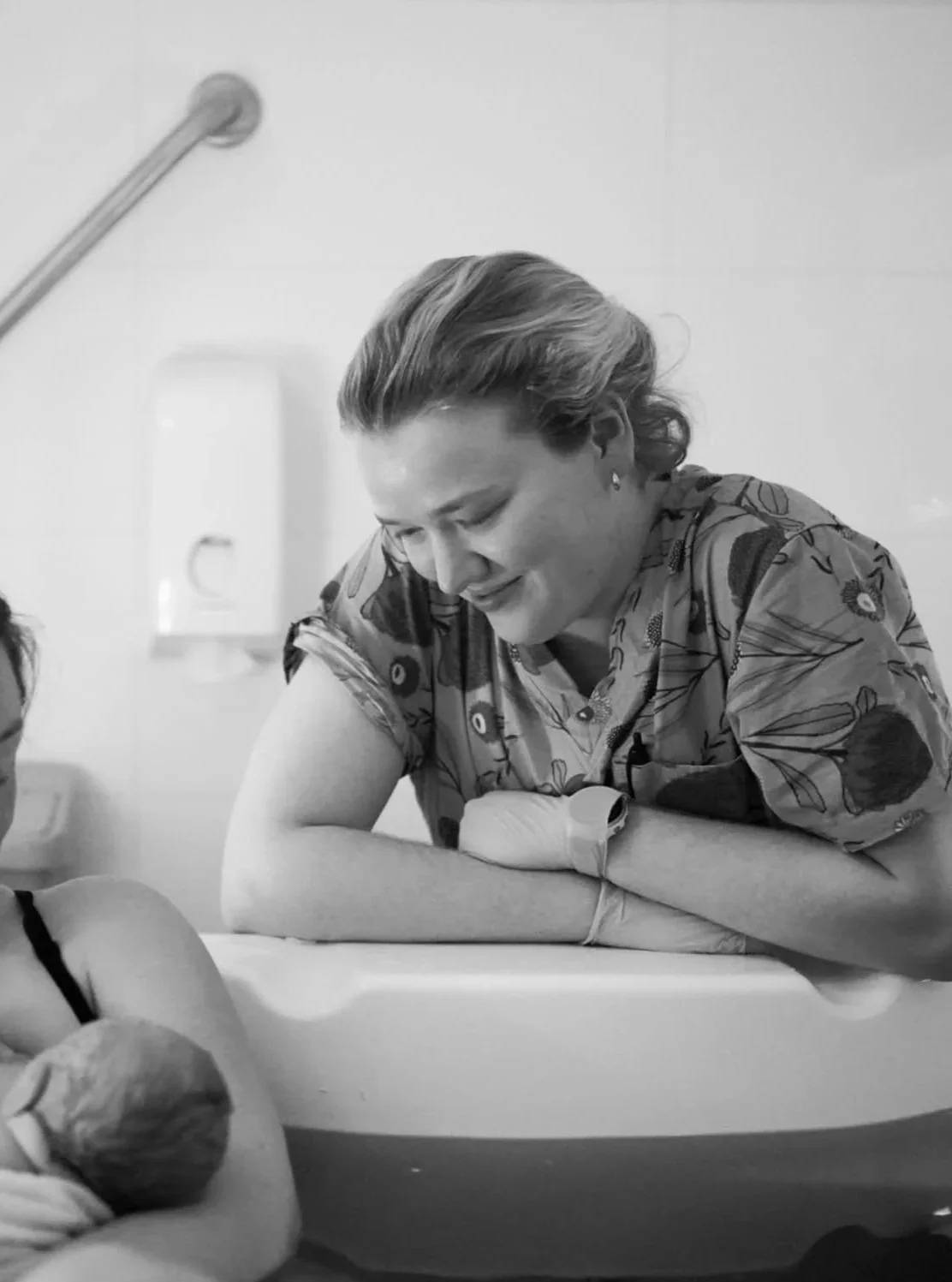 A woman holding a newborn baby in the bath, with her midwife looking on.