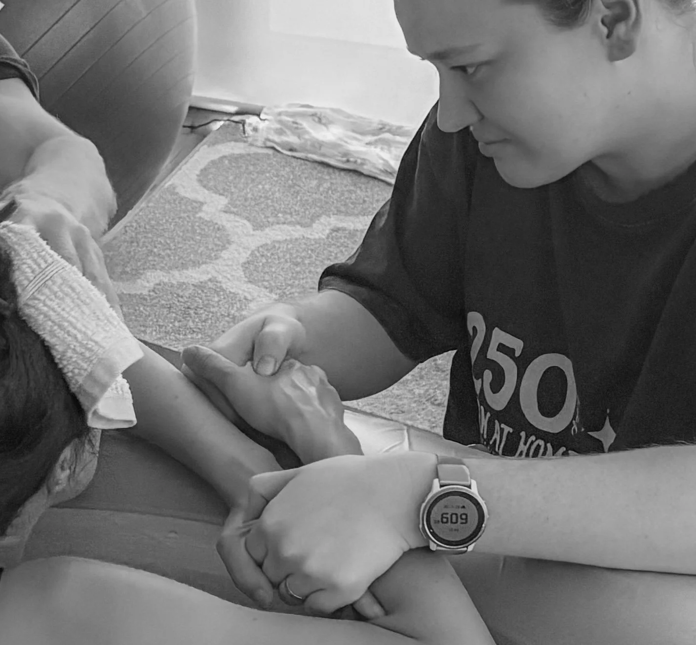 A woman is lying down with an arm extended, while a man is holding her hand and supporting her arm. The man appears to be giving a handshake or support in a caring manner.