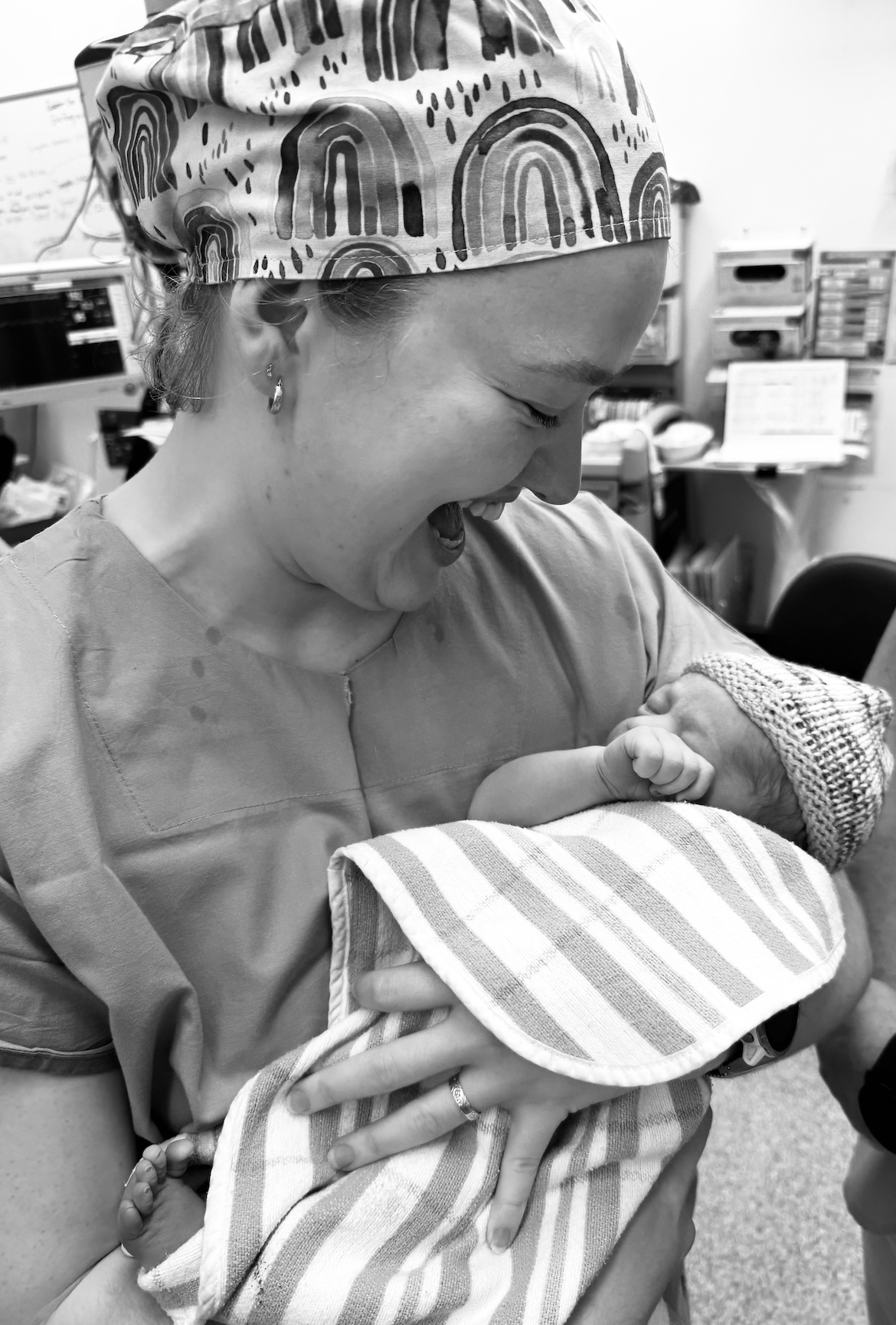 A woman in scrubs, wearing a patterned scrub cap, holding a newborn baby wrapped in a striped blanket and wearing a knit hat, in a hospital room.