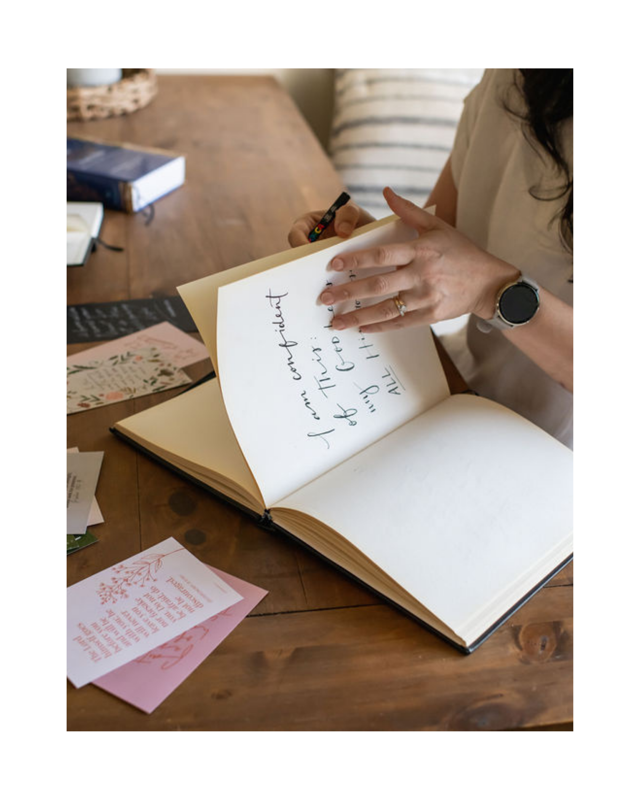 Picture of Jasmine Green writing in a blank artbook at a table, surrounded by her hand lettering Scripture art cards.