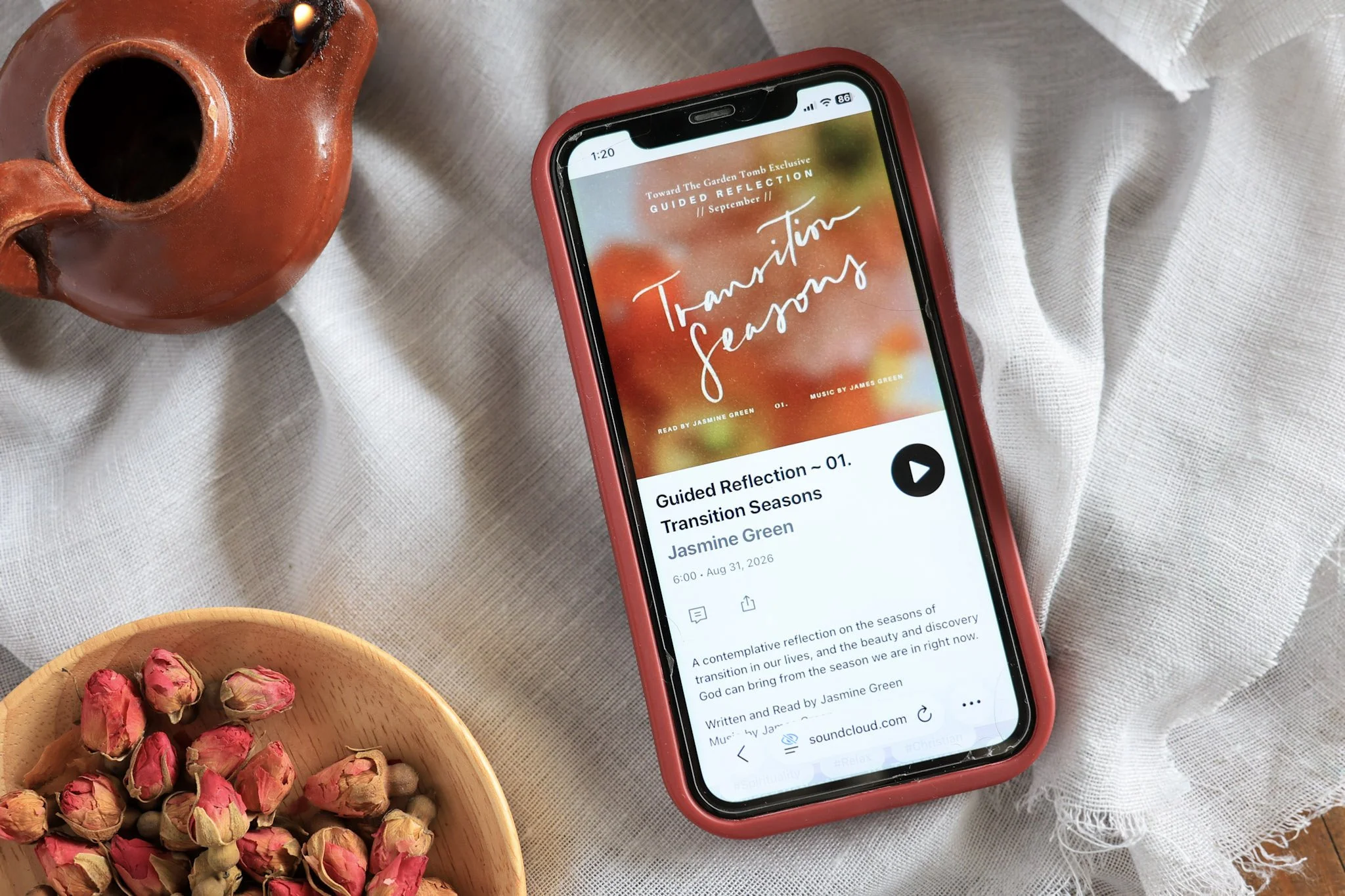 A smartphone with a red case displaying an audi app playing 'Transition Seasons' by Jasmine Green on a textured fabric surface, next to a wooden bowl filled with pink and green dried flower buds, and other small bowls nearby.