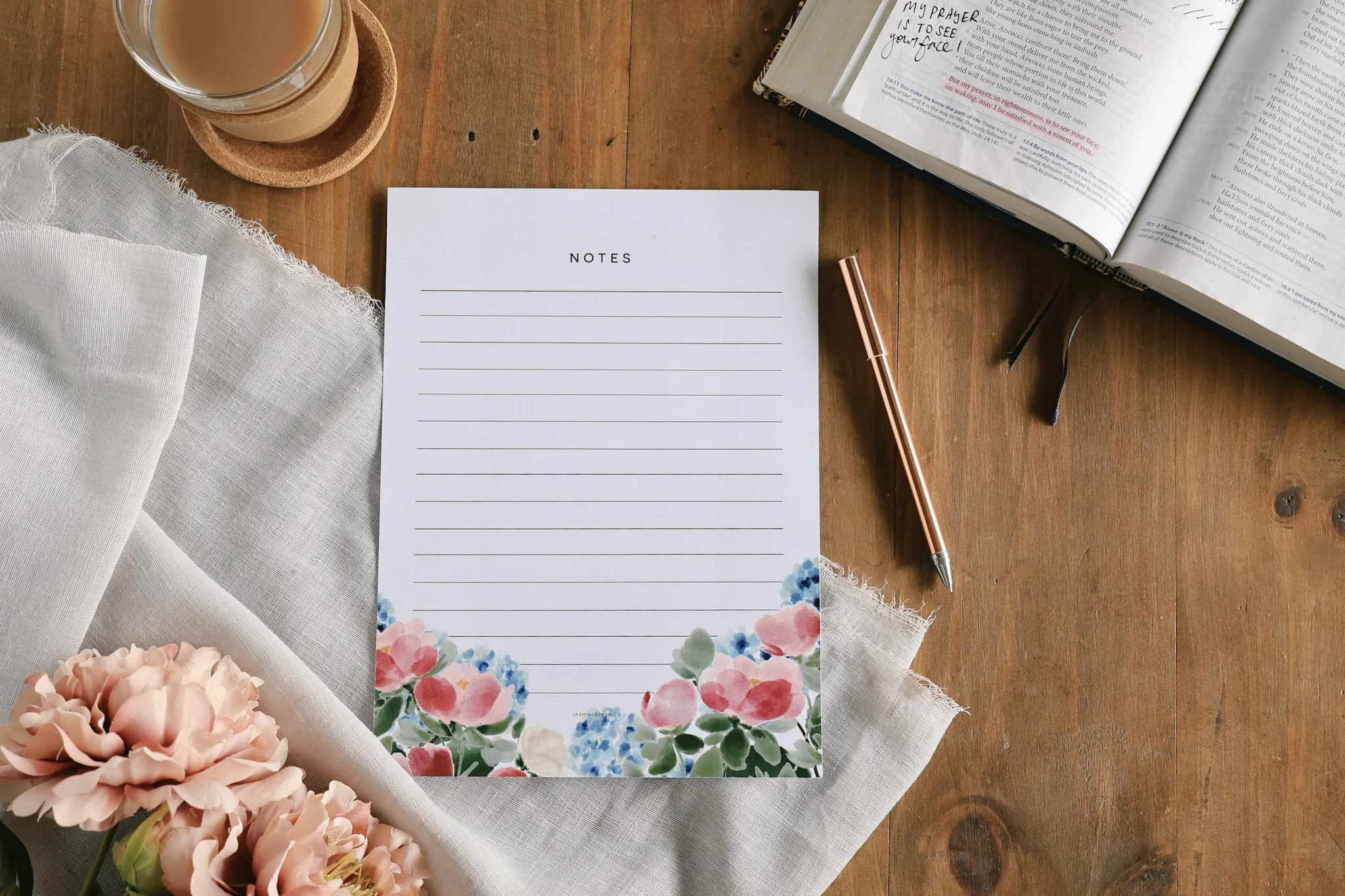 A wooden table holds a glass of tea on a cork coaster, an open Bible, a printed white notepad page with floral decoration, a pen, a piece of white fabric, and pink flowers.