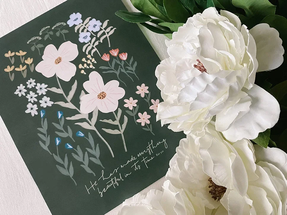 A dark green card with colorful illustrations of flowers and a handwritten quote, placed beside white peony flowers.