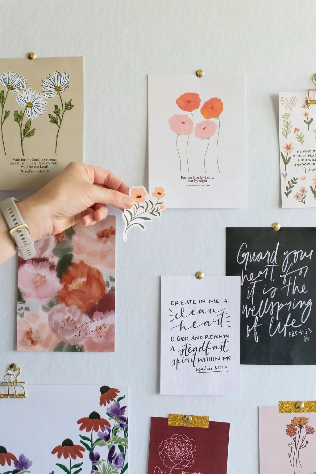 A collection of floral and inspirational quote cards pinned on a white wall, with a hand holding a flower-shaped sticker.