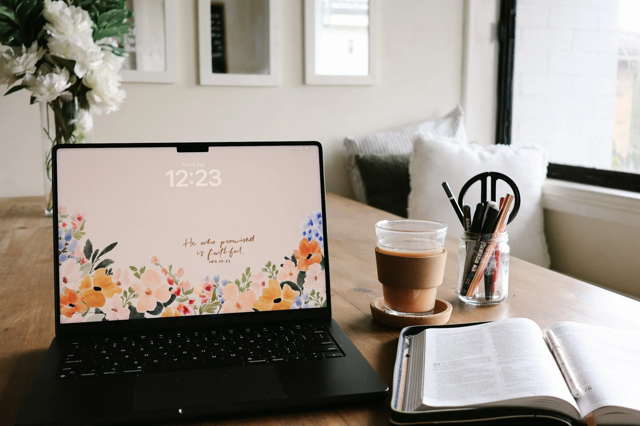 A laptop on a wooden desk displaying a floral background with a biblical quote, a notebook, a pen, a glass cup with pens, a cup of coffee, and a nearby open book. In the background, a window with light coming in, some pillows on a window seat, and wall decorations.