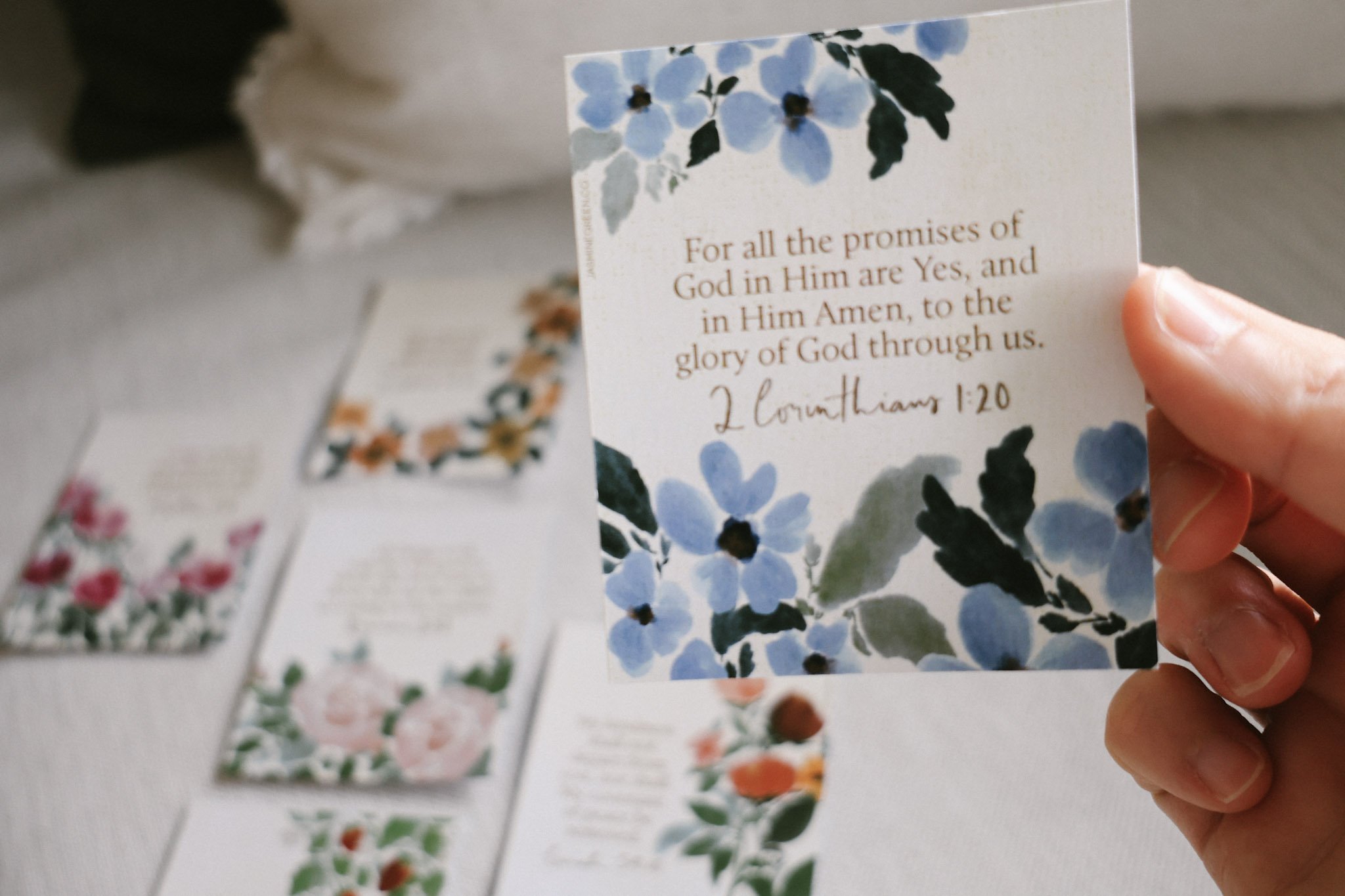 Close-up of a hand holding a small Scipture Art card with a floral design and a Bible verse, 2 Corinthians 1:20, printed on it.