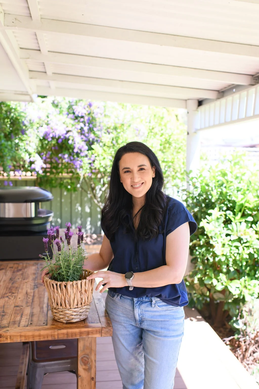 A woman with black hair, wearing a navy blue shirt and light blue jeans, standing outdoors on a porch with greenery and purple flowers in the background, smiling while holding a wicker basket of lavender flowers.