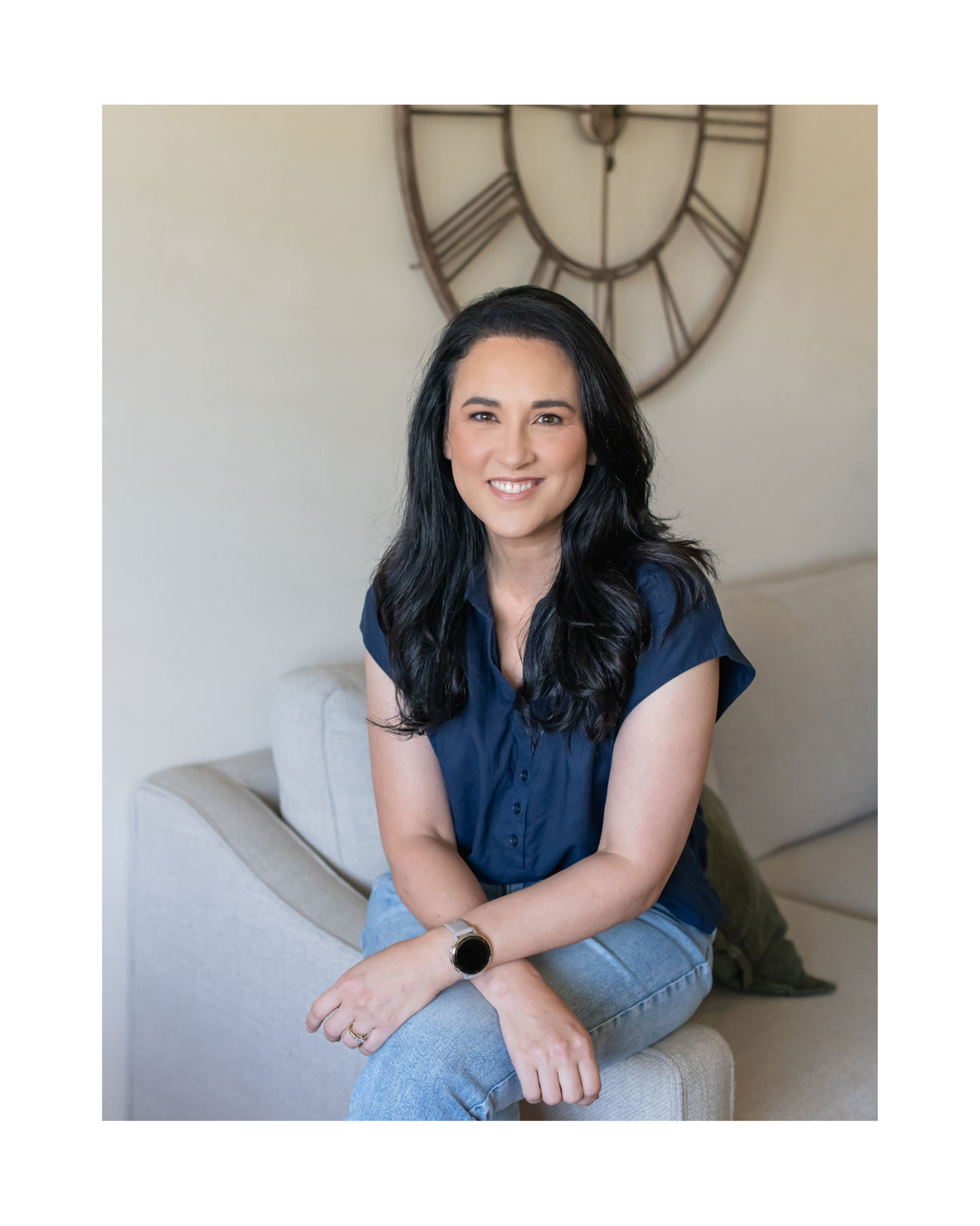 Christian artist and author, Jasmine Green, sits on a beige couch, wearing a blue button-up shirt and jeans, with her arms crossed and a watch on her left wrist, in front of a large round wall clock with Roman numerals.