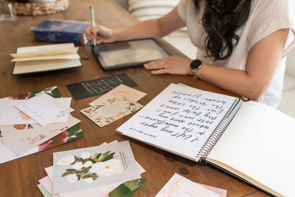 Person writing in a spiral notebook surrounded by greeting cards on a wooden table.