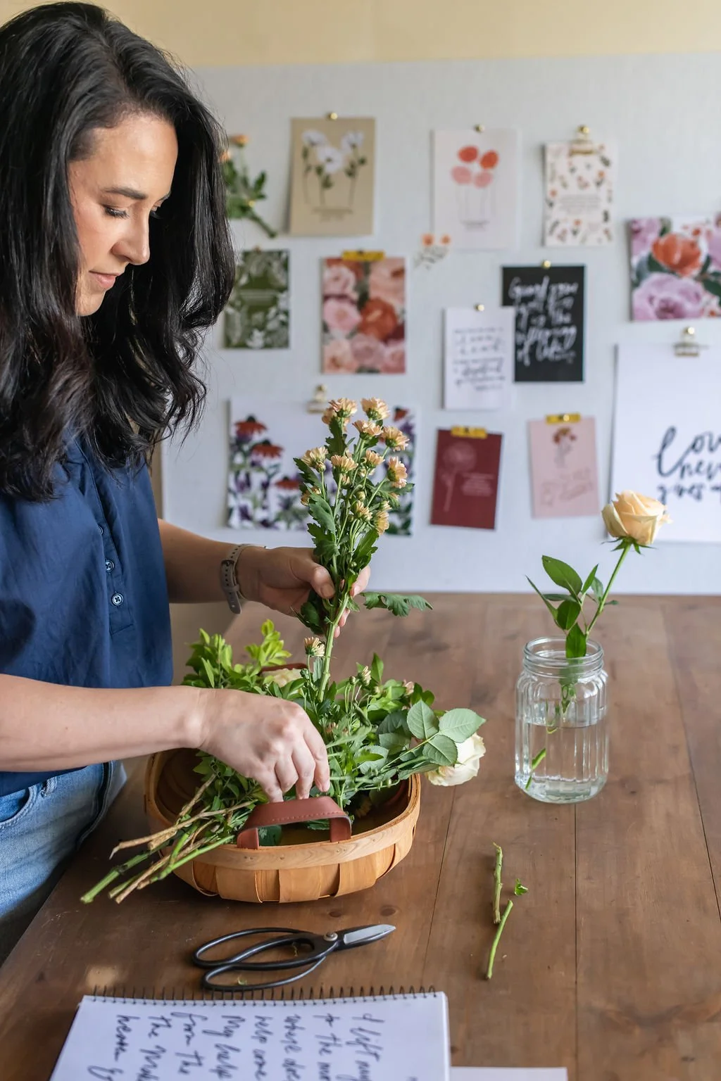 Woman arranging flowers in a basket at a wooden table with a mason jar of water and a single rose, with a wall of floral designs and quotes in the background.