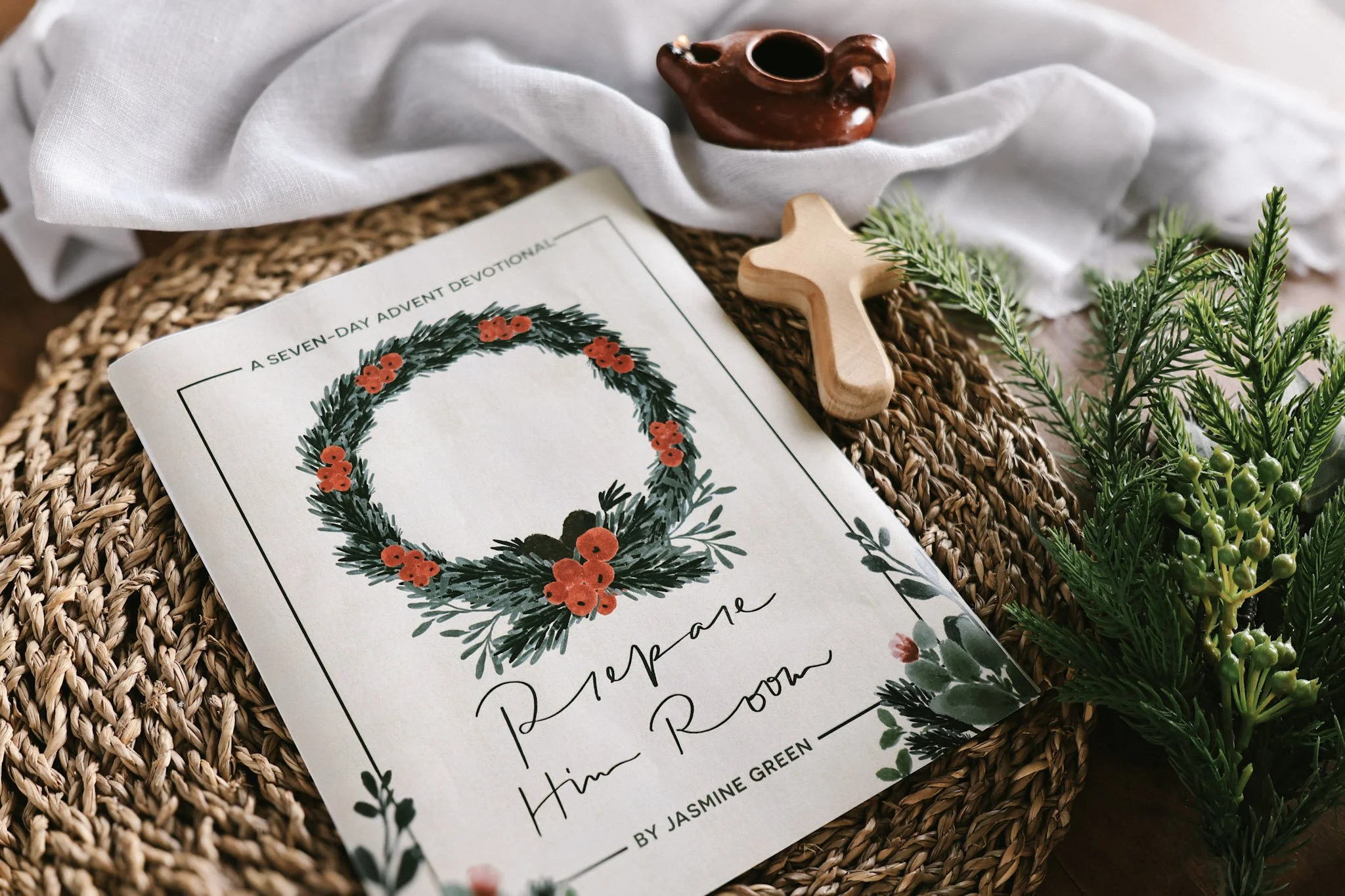 A printed Advent Devotional by Jasmine Green with a Christmas wreath illustration surrounded by greenery, placed on a woven surface, next to a small clay teapot and a wooden cross, with a white cloth partially covering the items.