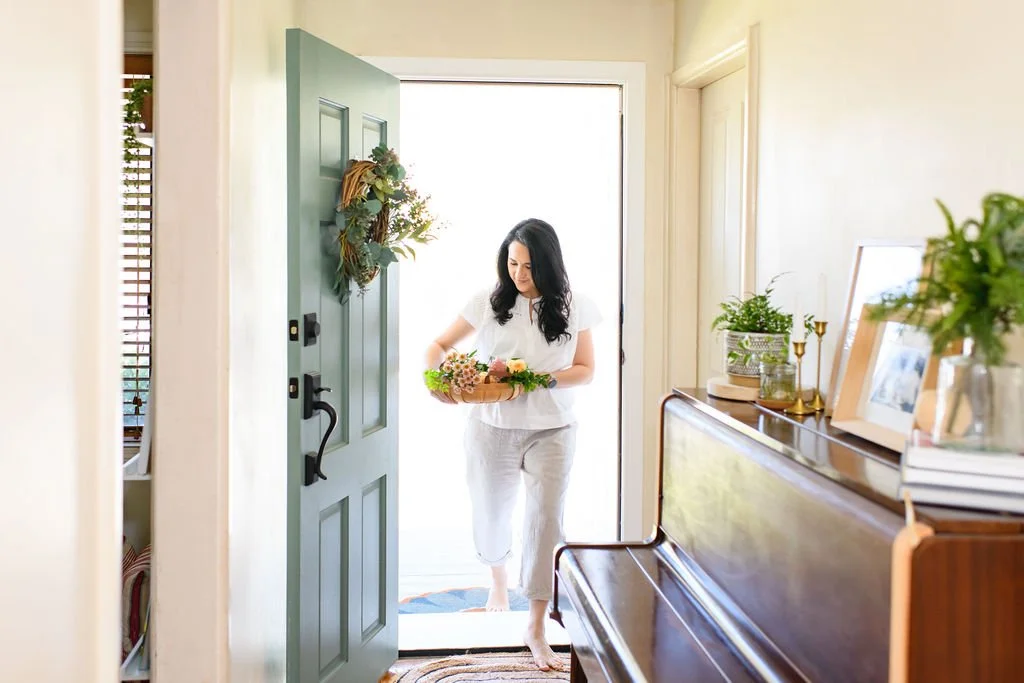 Woman at the door holding a basket of flowers in a brightly lit entryway.