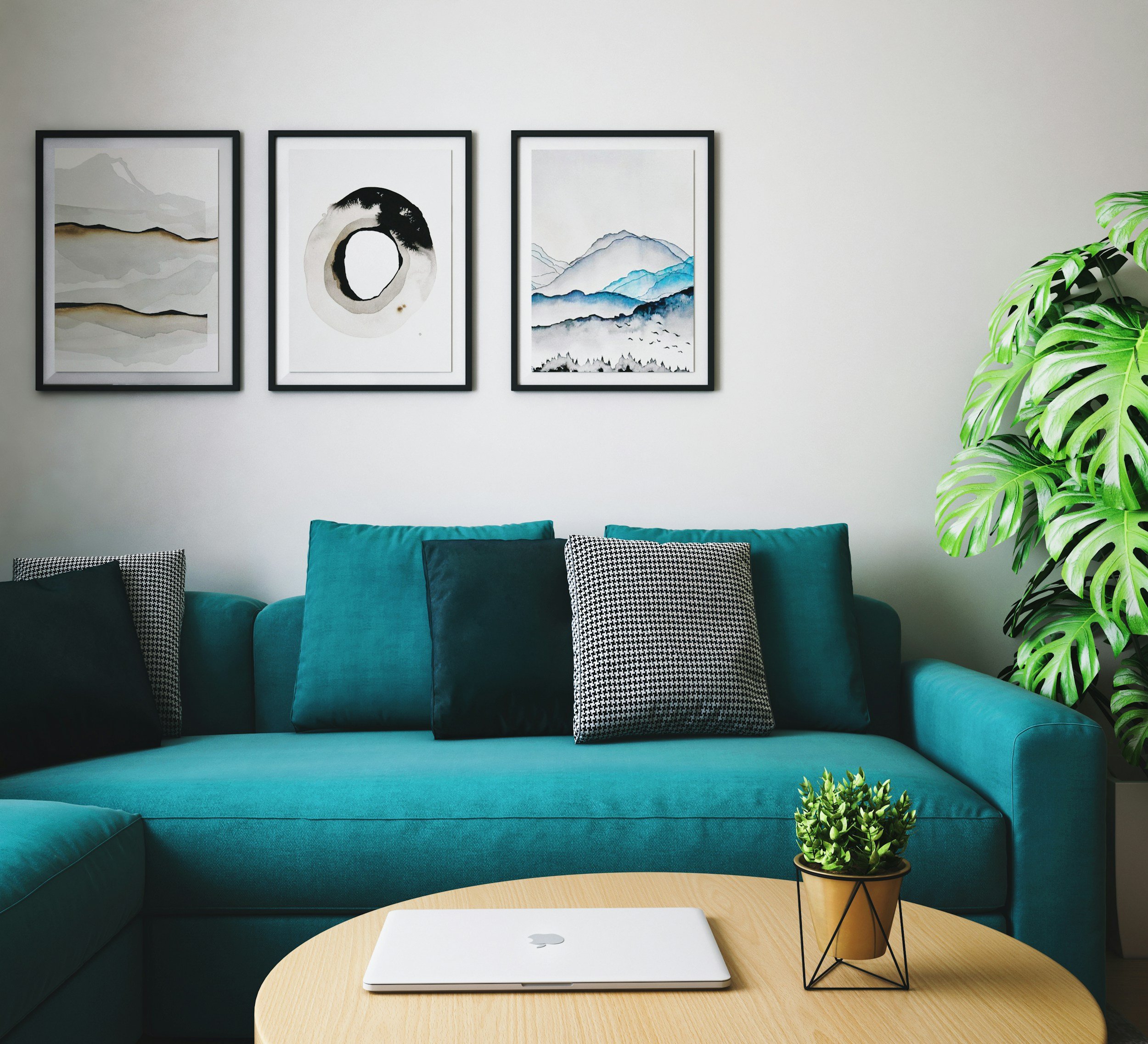 Living room with blue velvet sofa, three framed art pieces of abstract mountain and landscape scenes on the white wall, green plants on right side, wooden coffee table with a silver laptop, and patterned cushions on the sofa.