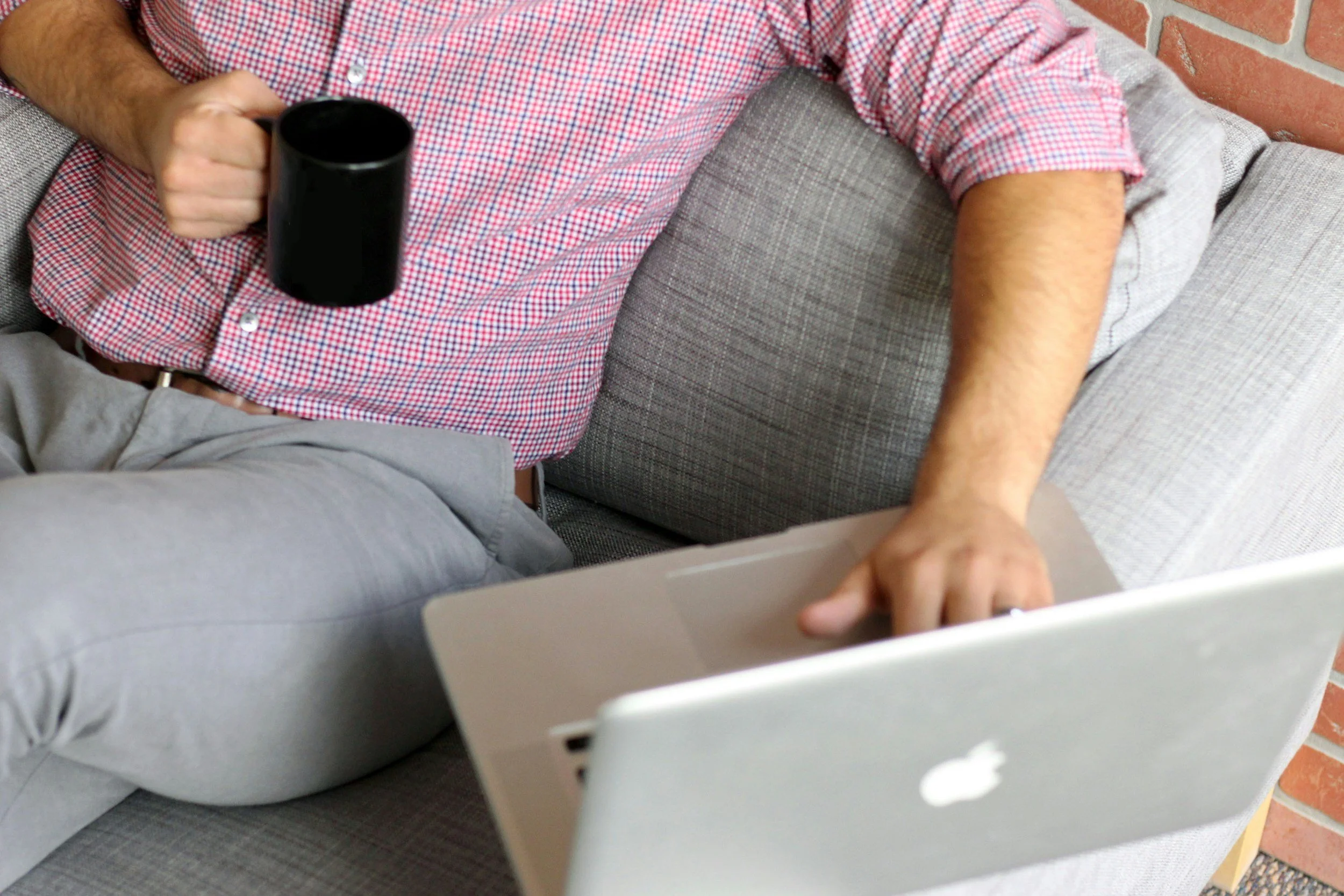 A person sitting on a couch, holding a black coffee mug in their right hand, wearing a red and white checkered shirt with sleeves rolled up. The person's left hand is resting on a white computer or tablet, and they are wearing light gray pants. The setting includes a gray couch with textured fabric and a brick wall background.